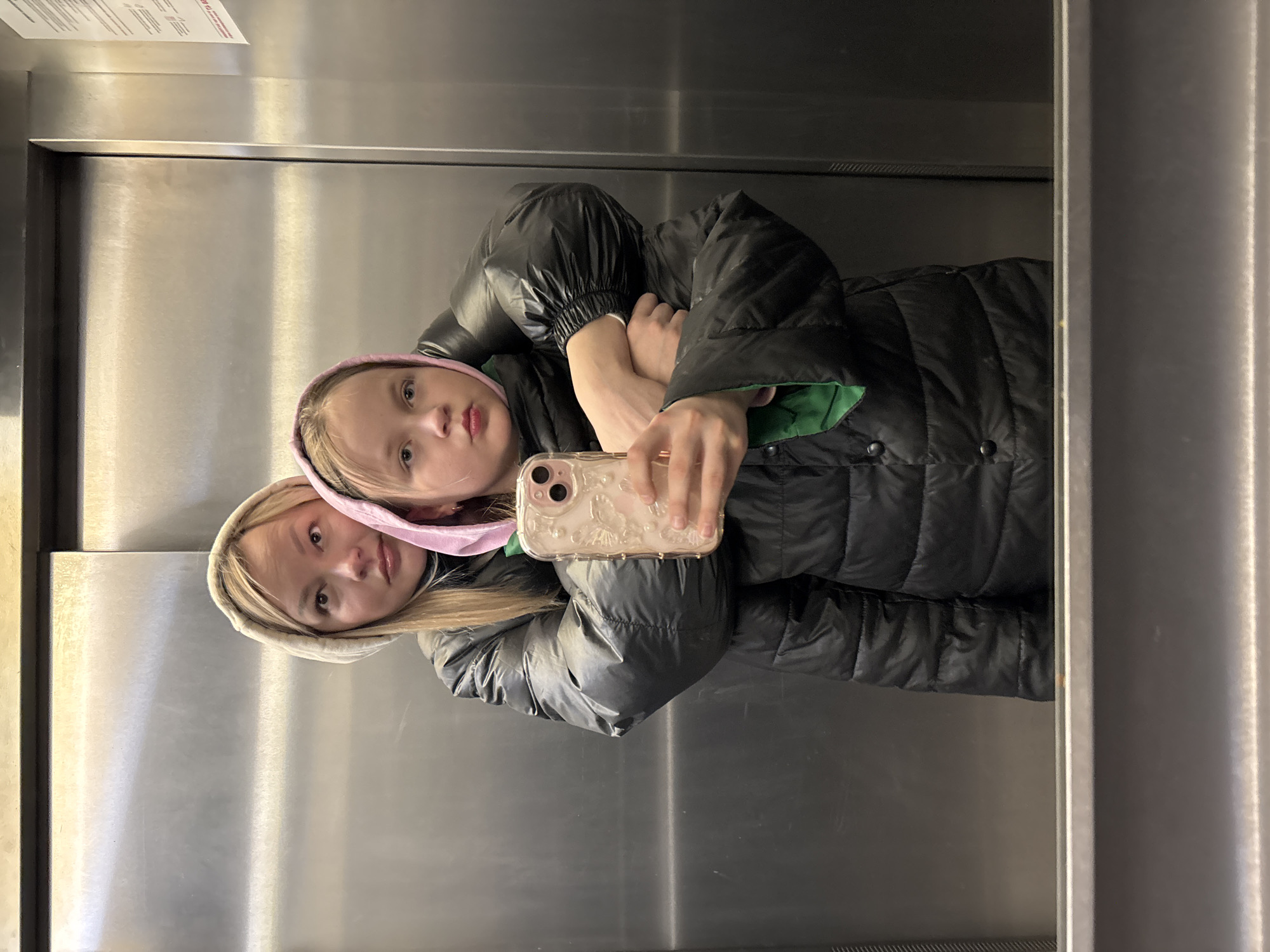 Two young blonde girls are taking a selfie in an elevator. The first girl is wearing a pink hooded sweatshirt and a black puffer coat with green lining. The second girl is wearing a cream-colored hooded sweatshirt and a light gray puffer coat. The elevator has stainless steel walls. There is a sign on the wall above the girls. The location is not specified, but it is in a city called Izhevsk, Russia.