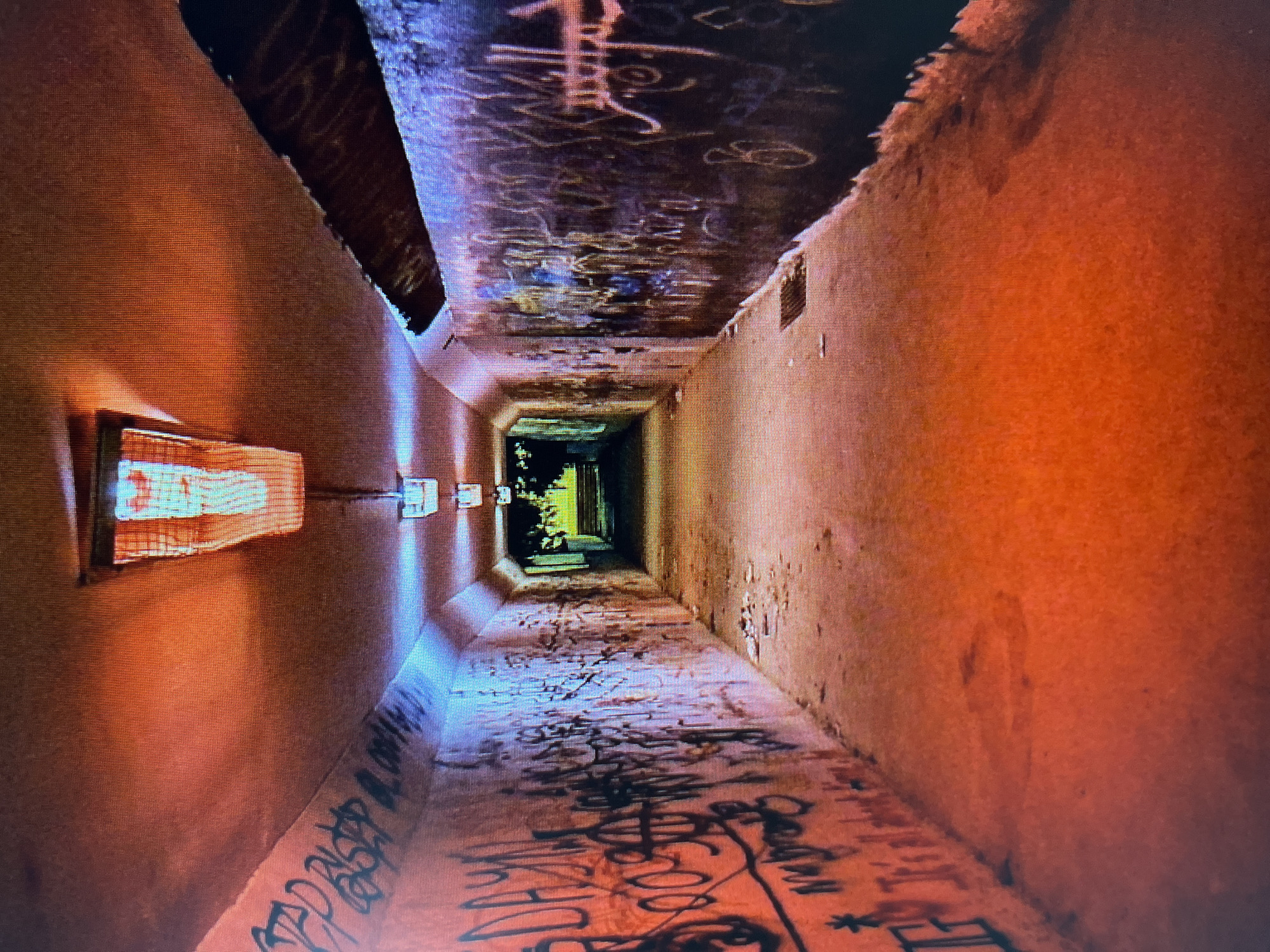 The media file depicts a long, narrow, and graffiti-covered concrete tunnel. The perspective is looking down the tunnel, creating a strong sense of depth and leading the viewer's eye towards a brightly lit opening at the far end. The tunnel walls and ceiling are covered in various forms of graffiti, primarily in black spray paint, with some lighter colored markings visible on the ceiling. The floor is also heavily marked with graffiti.
On the left wall, midway down the tunnel, is a rectangular light fixture with a translucent cover, emitting a warm, orange glow. There are also other, less distinct, square fixtures visible further down the tunnel on the left wall, also emitting light. The walls of the tunnel have a rough, textured surface and are illuminated by a warm, orange-red light that casts long shadows, suggesting either artificial lighting or a sunset. The overall mood is somewhat eerie and industrial, common to abandoned or underutilized urban spaces. The lighting creates a dramatic contrast between the shadowed interior and the bright exit, hinting at a passage or journey. No people are visible in the image. The time of day is indeterminate, but the lighting suggests it is either dawn or dusk, or that artificial lights are the primary illumination source.