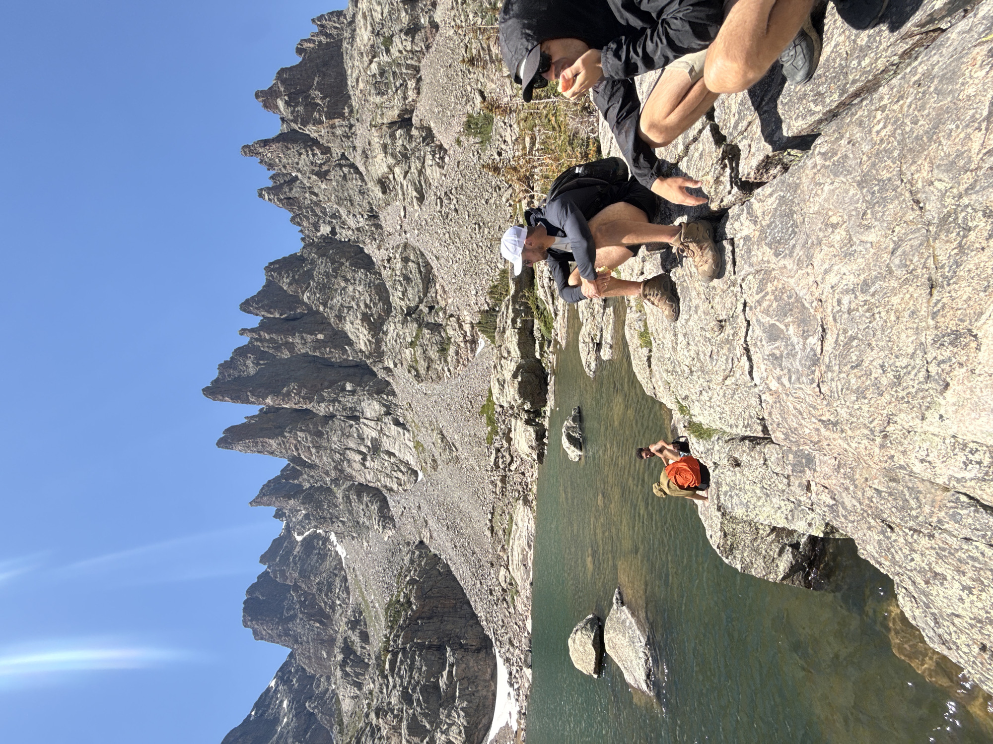 In Allenspark, Colorado, a scenic mountain landscape is depicted under a clear blue sky. In the foreground, two individuals are crouched on a rocky outcrop near a lake. The person on the left wears a white baseball cap, a dark long-sleeved shirt, and dark shorts with hiking boots. They are looking to the left. The person on the right is wearing a black hooded jacket and sunglasses, and is eating what appears to be a piece of fruit while looking towards the left. In the middle ground, near the water's edge, another individual is partially visible, wearing an orange backpack and dark shorts. Behind them, a tranquil lake reflects the rugged, rocky mountains that dominate the background. The mountains are characterized by sharp, jagged peaks, with patches of snow and scree slopes visible on their surfaces. The overall scene conveys a sense of outdoor adventure and natural beauty in a mountainous region.