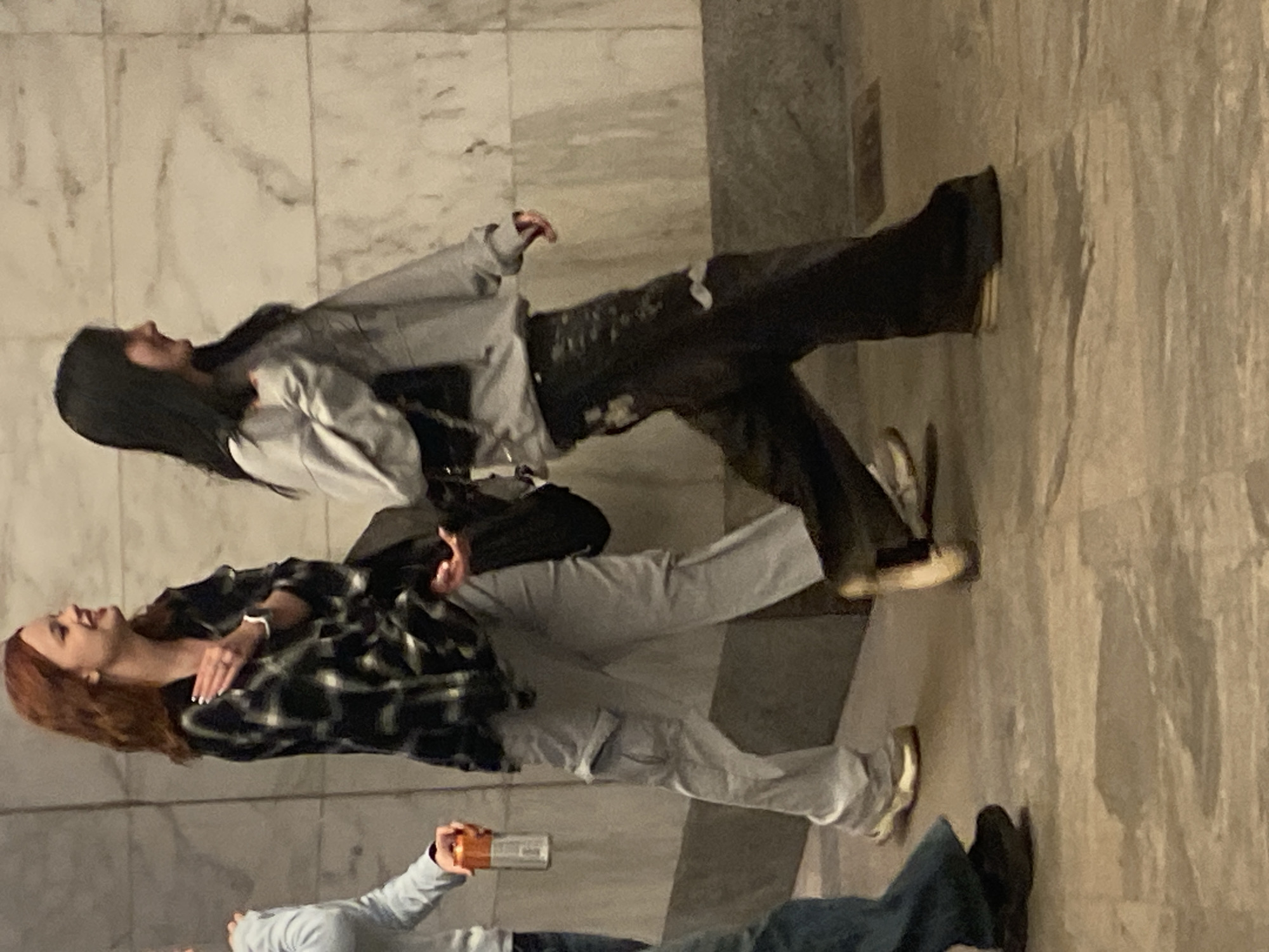 Three individuals are walking side-by-side in an indoor setting, possibly a building with marble walls. The person on the left, partially visible, is wearing a light blue long-sleeved shirt and blue jeans, holding an orange bottle. The person in the center has reddish-brown hair and is wearing a black and white plaid shirt over light gray cargo pants and beige sneakers. They are looking to their left and appear to be speaking. The person on the right has long black hair and is wearing a light gray hoodie with dark, distressed pants and dark chunky shoes. They are carrying a black bag over their shoulder and looking forward. The ground is paved with light-colored tiles. The overall lighting is dim. There is no information in the image to confirm or deny the location in Khimki, Russia.