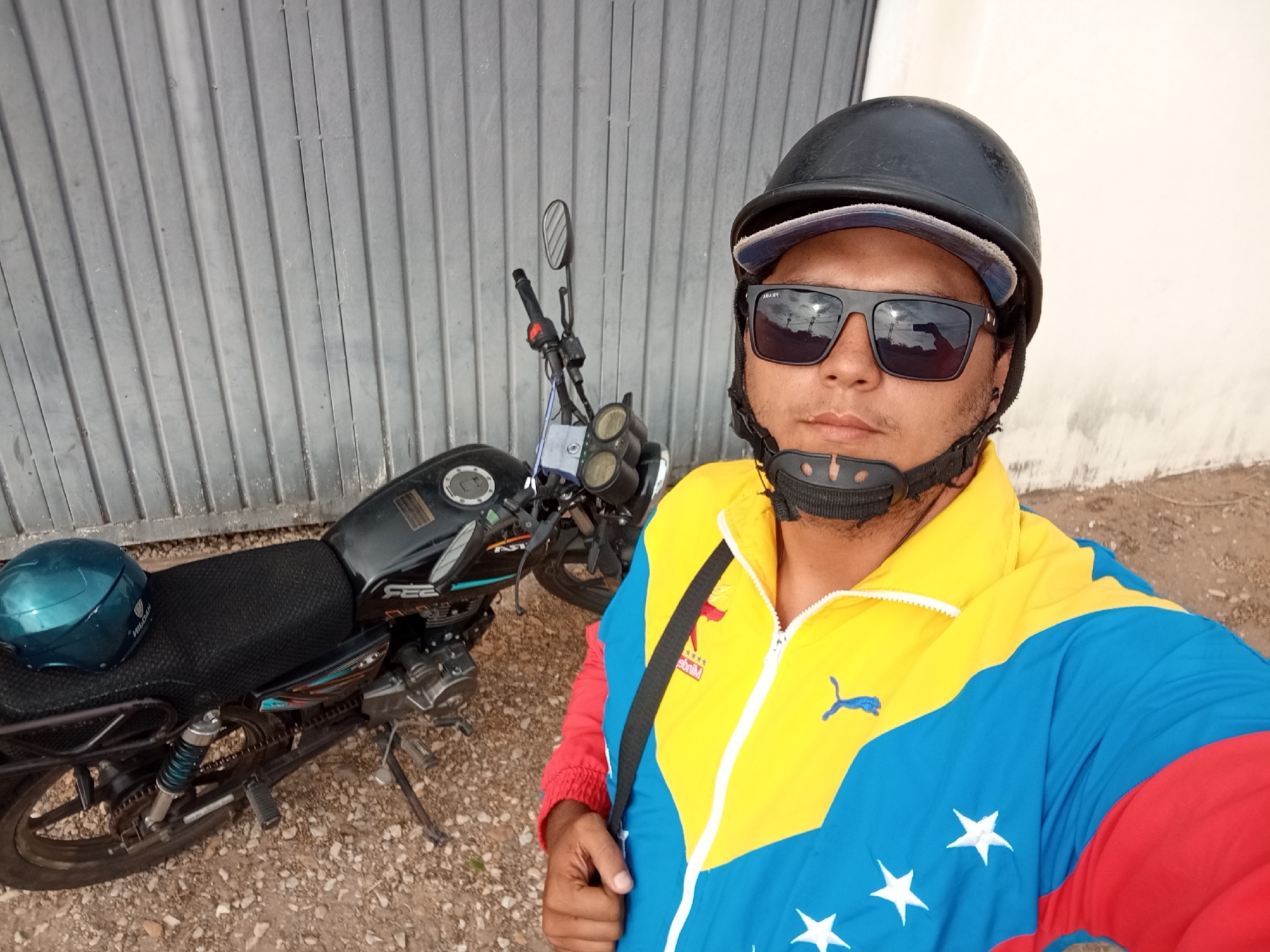 A medium shot captures a man in front of a gray metal garage door next to his parked black motorcycle. The man wears a black motorcycle helmet with a gray-brimmed hat underneath, dark sunglasses, and a yellow, blue, and red jacket with white stars. The motorcycle, parked on a gravel surface, has a blue helmet placed on its seat. The location is Juan Griego, Venezuela.