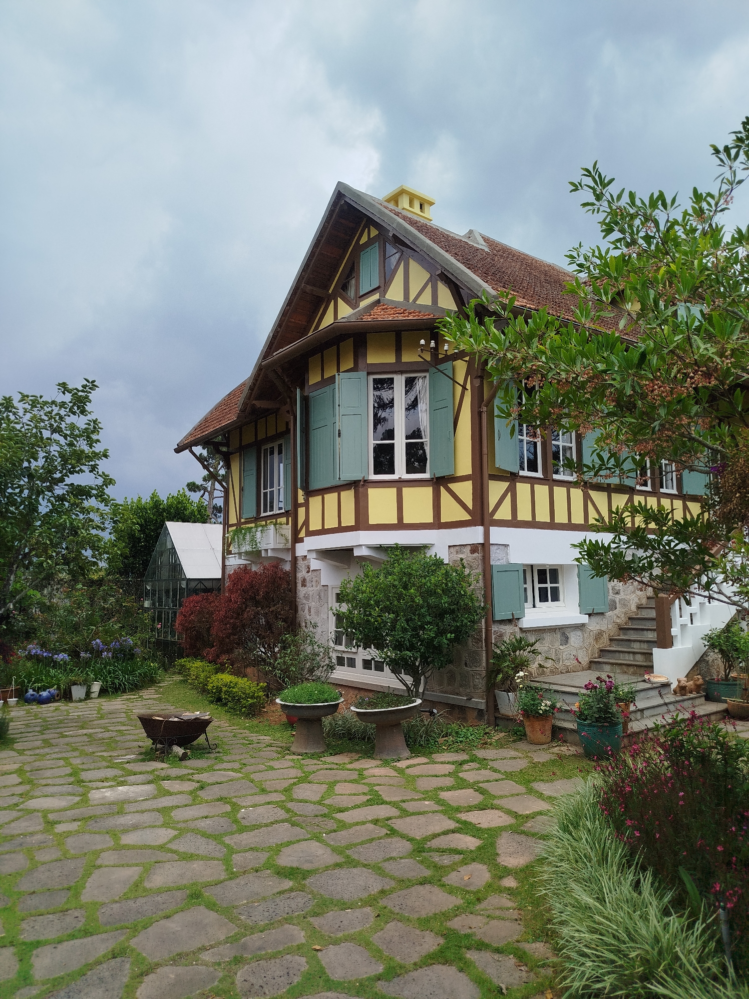 The image depicts a Tudor-style house in Da Lat, Vietnam. The house has a yellow and white facade with dark brown timber framing, green shutters, and a red-tiled roof. There is a small greenhouse attached to the side of the house. A stone pathway leads to the front of the house, with various plants and flowers lining the pathway. The sky is overcast. There are no people visible in the image.