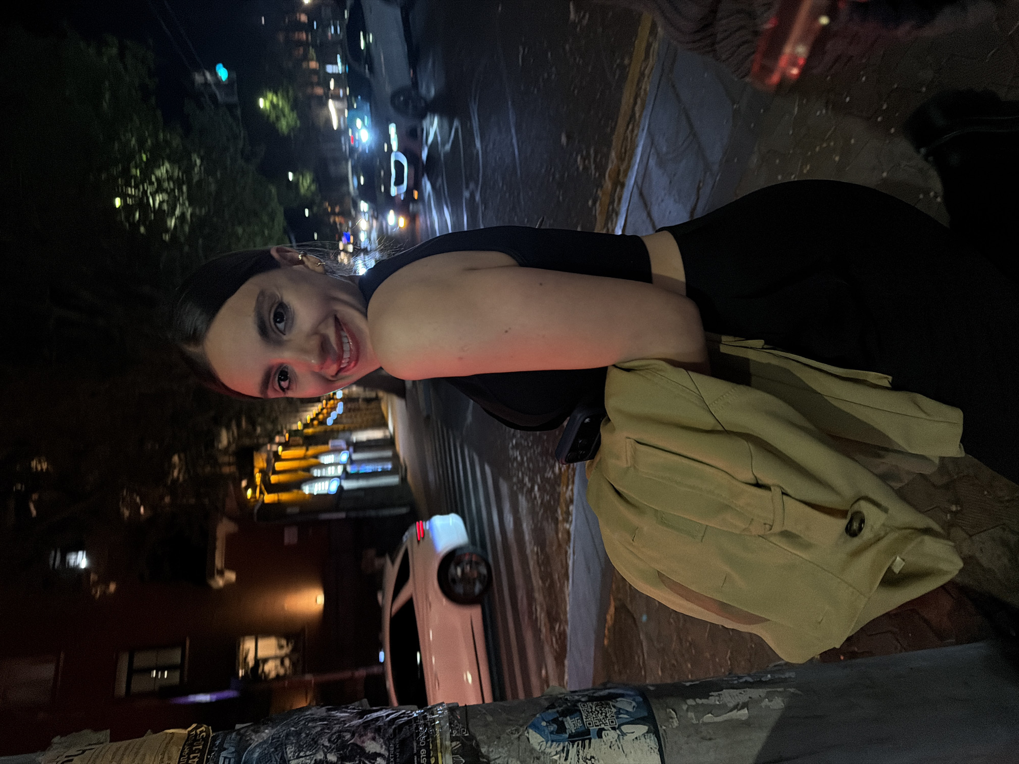 A young woman poses for a photo on a city street at night. She wears a black top and pants, a light brown jacket is draped over her arm. In the background, street lights illuminate buildings and parked cars. Based on the urban environment and the presence of Armenian script on a sign near the bottom of the frame, this photo appears to be taken in Yerevan, Armenia.