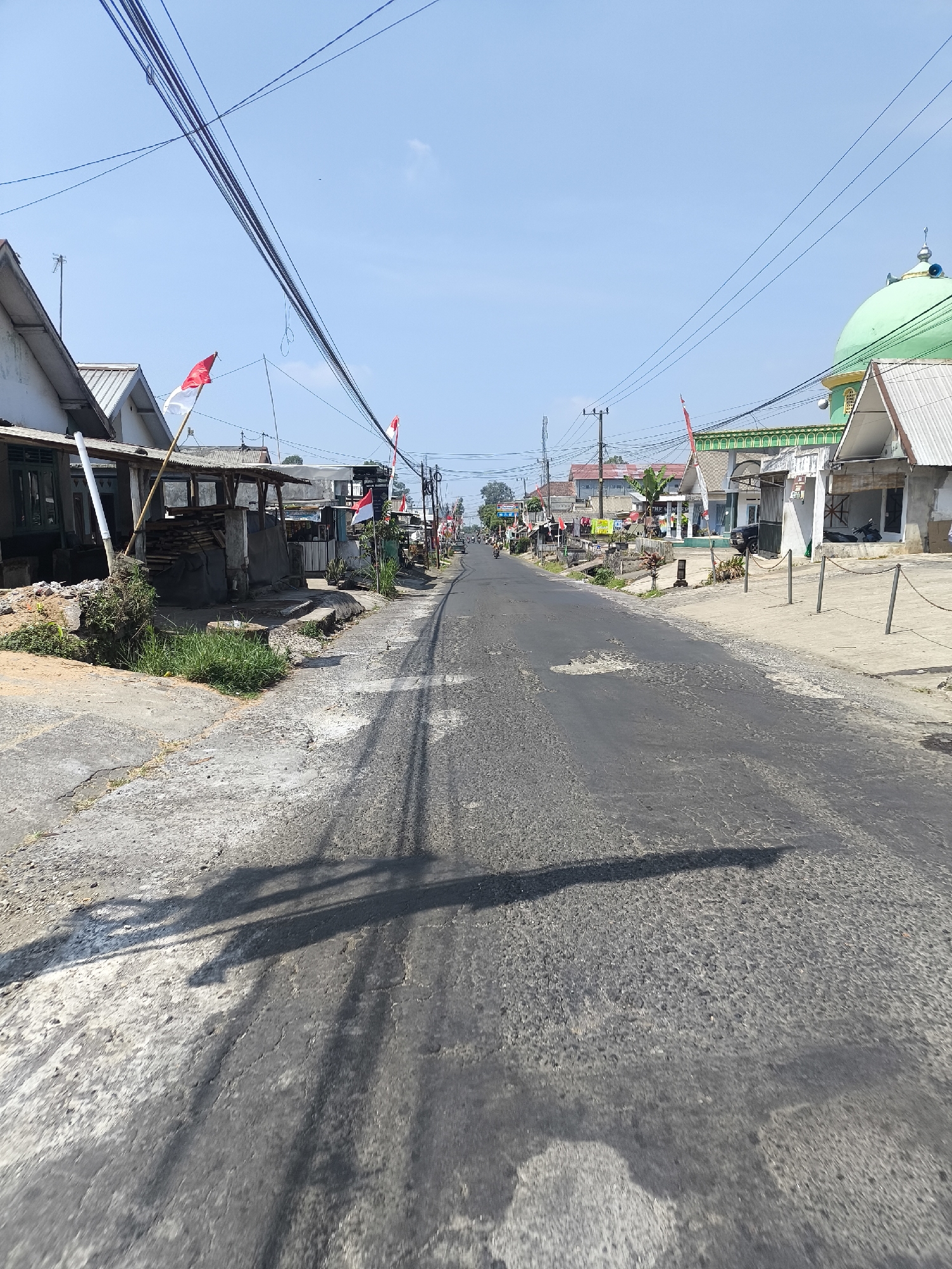 The image depicts a street scene in Senduro, Indonesia, under a clear blue sky. The road surface is asphalt, showing signs of wear with patches and cracks. On either side of the road, there are buildings, some with white facades and sloping roofs, and others appearing more rustic with wooden structures and corrugated metal roofs. Red and white Indonesian flags are displayed in several locations, indicating a patriotic or celebratory occasion. A prominent green-domed building, likely a mosque, is visible on the right side of the street. Power lines crisscross overhead. The scene is largely devoid of people, though a few vehicles, including motorcycles and a car, are present in the distance. Shadows of the viewer and overhead objects are cast on the road. The overall environment suggests a typical day in a semi-urban or rural Indonesian setting.