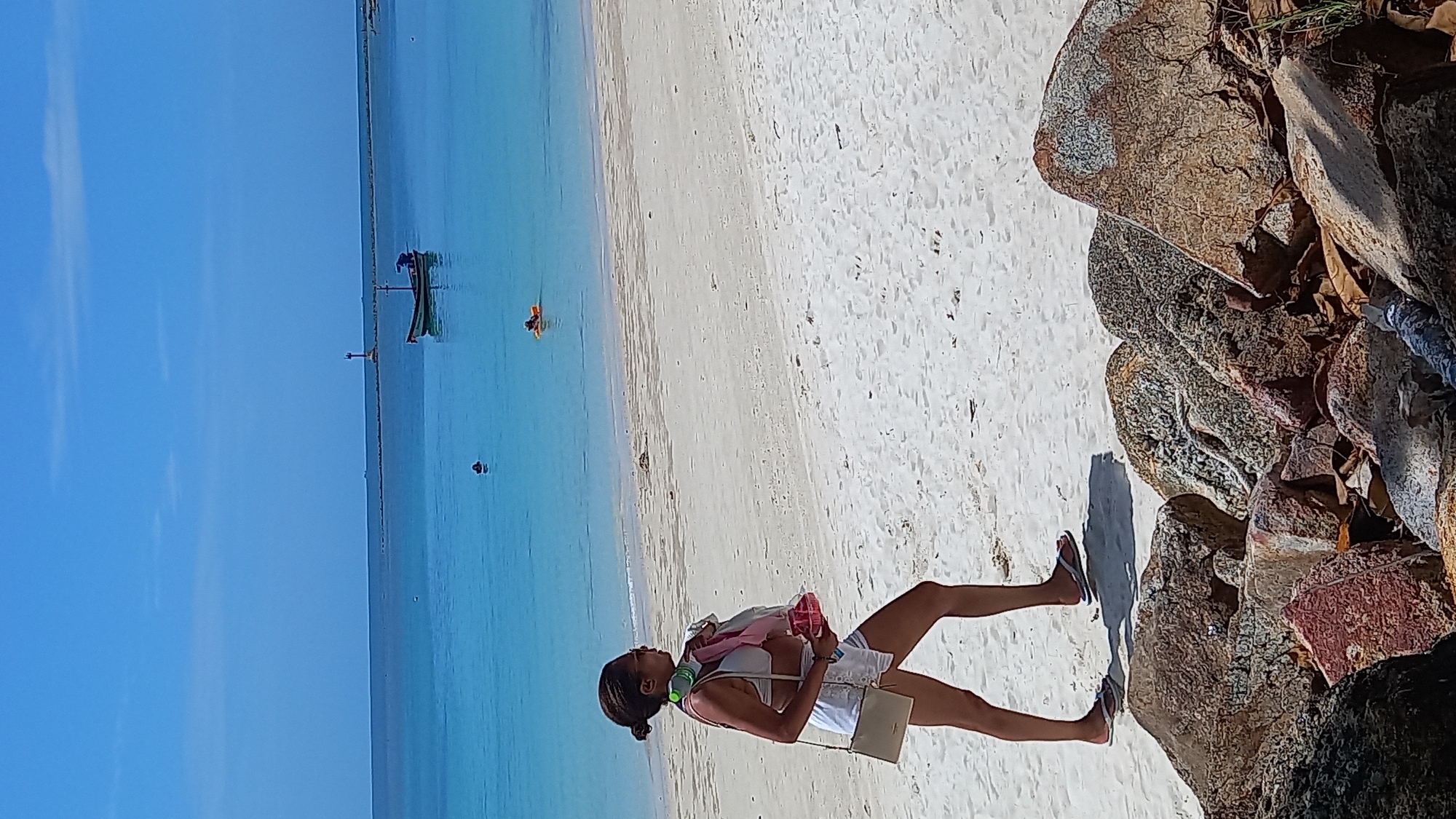 A person walks on a sandy beach in the Ko Pha-ngan District, Thailand, next to large rocks. The person is wearing white shorts and a white top, with a beige purse slung over their shoulder and headphones around their neck. They are carrying a pink object and a red container. The ocean is a bright turquoise, with a small boat and a lighthouse visible in the distance. The sky is clear and blue.
