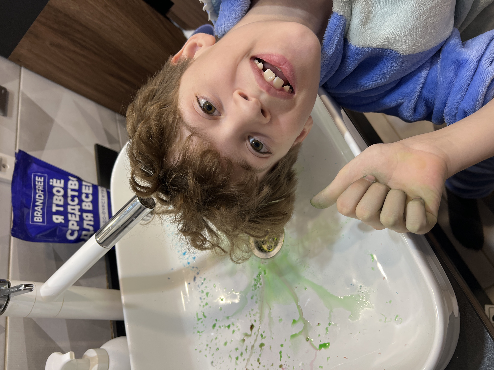 A boy, facing the camera, has his head upside down over a bathroom sink. He is fair-skinned with light brown, curly hair and is wearing a blue and white robe. He is pointing with his right hand towards the sink. Green and blue liquid and granules are scattered inside the white ceramic sink. A silver faucet is visible on the left side of the sink, along with a blue package labeled "BRANDFREE Я ТВОЁ СРЕДСТВО ДЛЯ ВСЕГО!". The background includes a brown cabinet above and white tiled walls. The image suggests a playful or mischievous interaction with a cleaning product in a bathroom setting. No information about the city of Noviy, Russia is present.