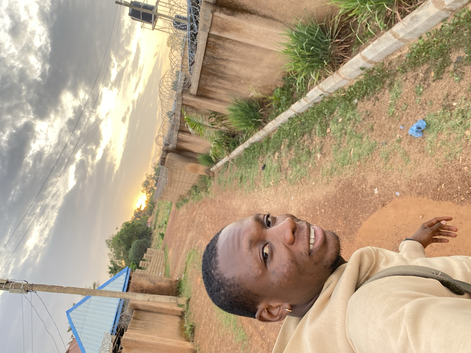 This vertical outdoor image captures a young Black man smiling directly at the camera, set against a backdrop of a dirt path and various structures, likely in Bukuru, Nigeria.
The primary subject is a man in the foreground, positioned in the lower left portion of the frame. He has short, dark hair, a small gold hoop earring in his left ear, and is wearing a light tan/beige hoodie or long-sleeved top, with a dark strap across his chest. He appears happy, with a broad smile revealing his teeth.
The scene unfolds under a dramatic, partly cloudy sky, with a bright golden glow from the low sun, indicating either sunrise or sunset. The ground is a reddish-brown dirt path, interspersed with patches of green grass and weeds. In the mid-right, a small blue plastic object lies on the dirt. A concrete wall topped with coiled razor wire extends along the right side of the path, with overgrown grass and bushes behind it. Further back on the right, a black water tank rests on a metal stand. On the left, a utility pole with wires runs vertically, and behind it, a building with a light blue corrugated metal roof is visible. Another wall, constructed from concrete blocks, can be seen in the far left background. The overall setting suggests a practical, possibly developing, outdoor environment. No visible text is present in the image.