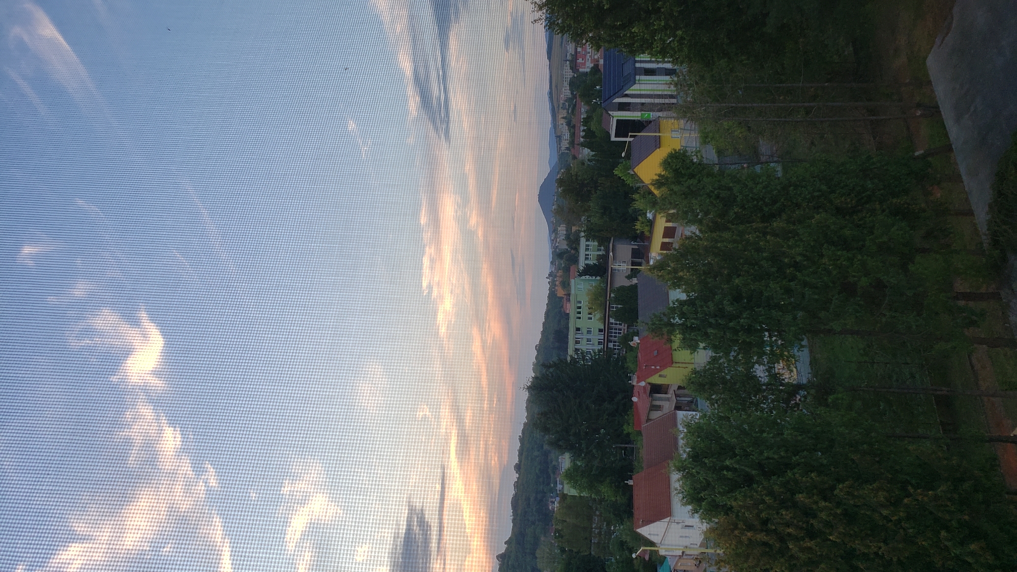 This is a photograph of a sunset over a residential area in Prešov, Slovakia, taken through a window screen. The sky is a gradient of blue, with soft pink and orange clouds scattered across it, indicating the sun is low on the horizon. Below the sky, a landscape of trees, houses, and buildings is visible. The houses in the foreground have red and yellow roofs. Further back, there are apartment buildings and a hill in the distance. The presence of a window screen is evident in the grid pattern overlaying the entire image.
