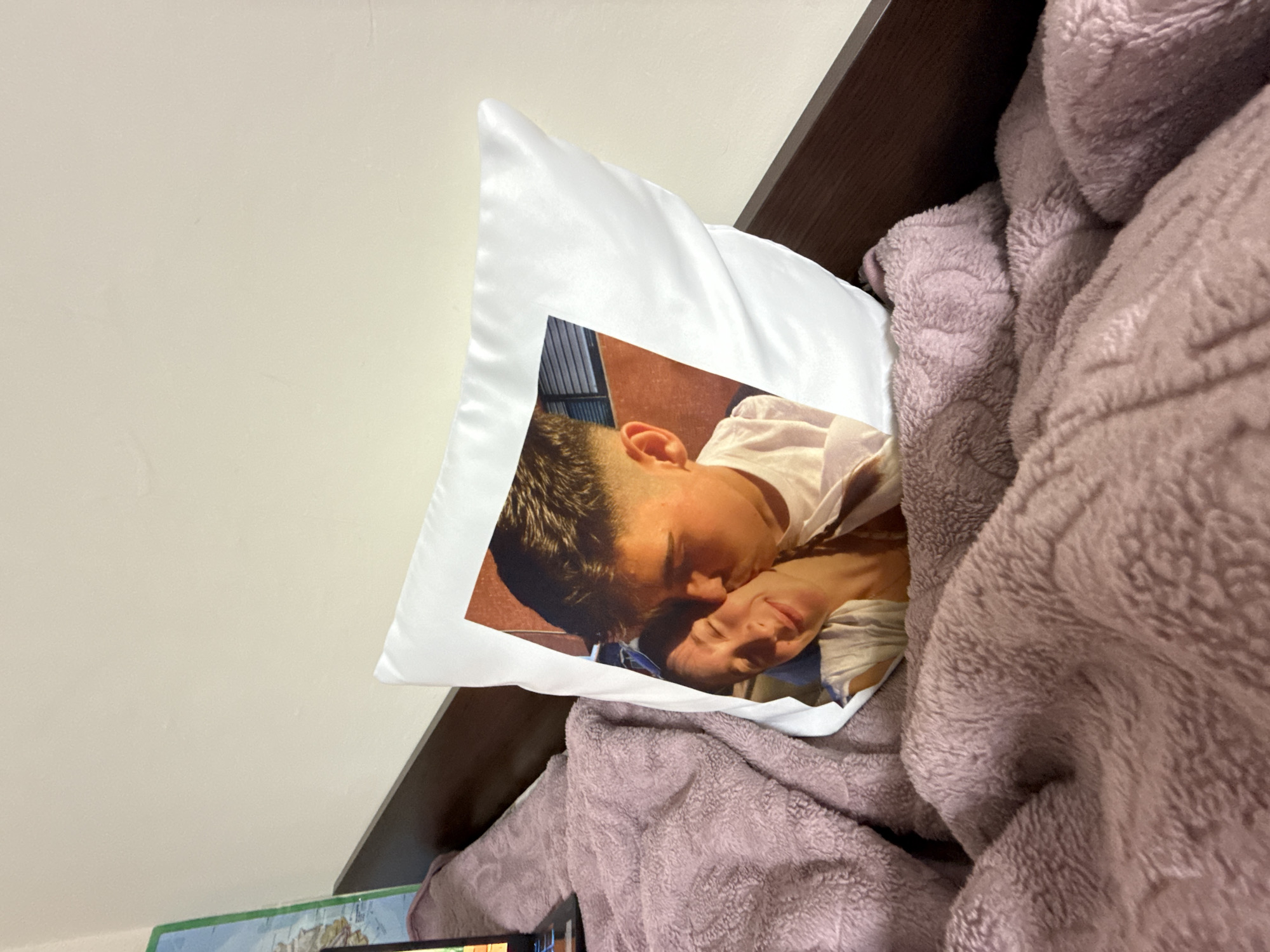 A close-up shot shows a white pillow with a printed image of two people lying on a bed. The bed has a brown headboard and is covered with a light purple, textured blanket. The setting appears to be indoors. No information is present in the image that allows for the identification of a specific location, including Sofia, Bulgaria.
