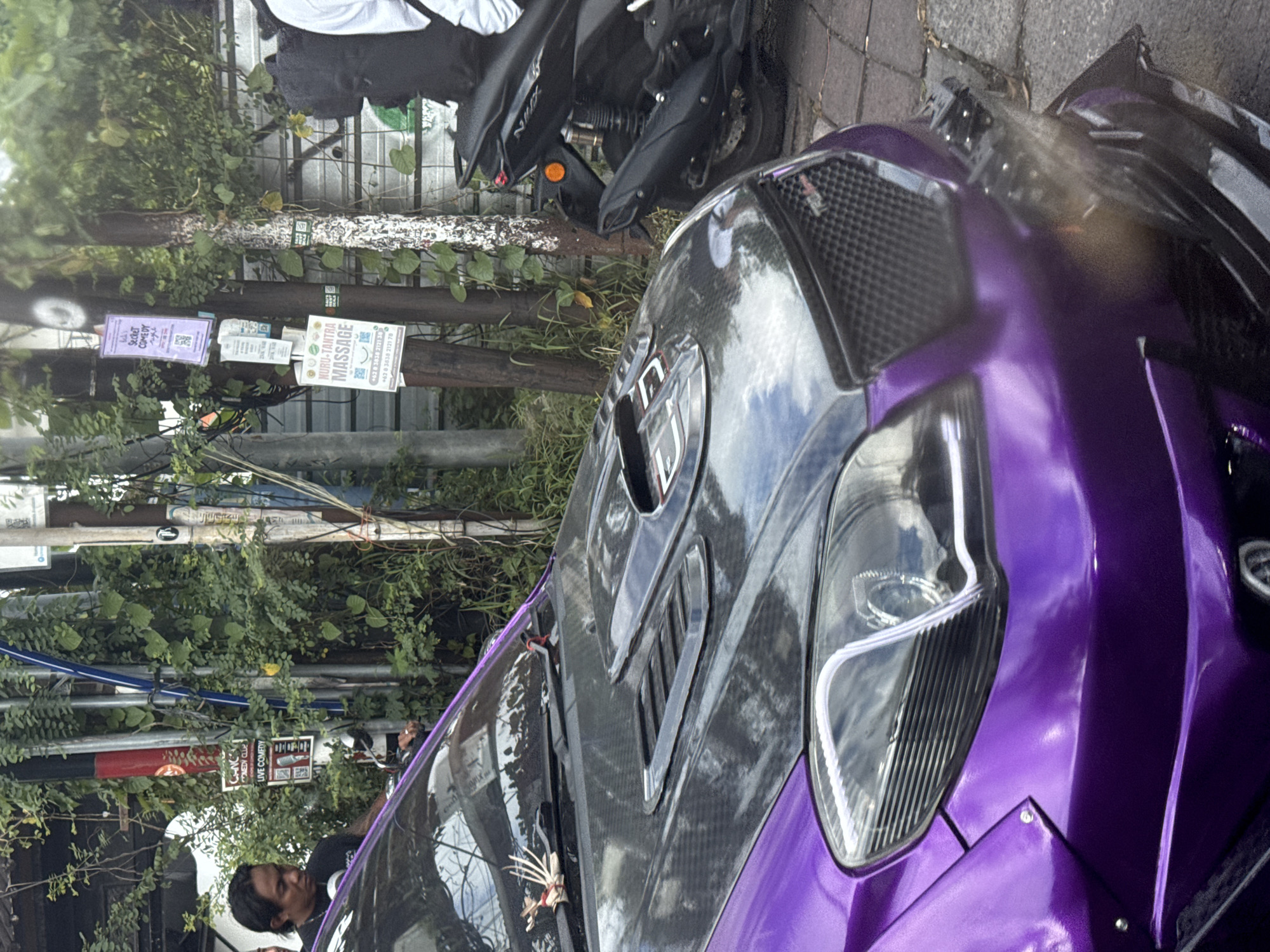 The image captures an outdoor, street-side scene, presented in a rotated orientation, depicting a customized purple car, a scooter, and lush vegetation.
The primary subject is a striking metallic purple car, likely a highly modified sports car or a custom build. It features a prominent carbon fiber hood with aggressive vents and scoops, along with modern projector-style headlights. A small, decorative charm resembling bones or spikes is visible near the car's windshield. Behind the purple car, partially visible, is a black scooter, identifiable as a Yamaha NMAX. A person with dark hair can be seen in the lower-left corner of the frame and reflected on the car's surface, seemingly observing the vehicle or the scene.
The setting appears to be an urban or semi-urban environment, characterized by abundant green foliage, including trees, vines, and bushes, which heavily line the background and foreground. Utility poles or posts, some with an overgrown, dirt-streaked wooden railing, are visible and serve as an attachment point for various advertisements. The ground, visible on the right side of the rotated image, is paved with stones or concrete.
Several flyers and posters are attached to the wooden posts. Visible text includes: a purple poster advertising "Secret COMEDY night," another poster for "CANC COMEDY CLUE LIVE COMEDY," and a larger white poster for "NURU-TANTRA MASSAGE" with Indonesian phone numbers: "+62 83838 2121 38" and "+62 0 3638 2121 78". The lighting suggests it is daytime, with diffused light, possibly indicating an overcast sky. The Indonesian phone numbers and lush greenery are consistent with the provided location context of North Kuta, Indonesia.