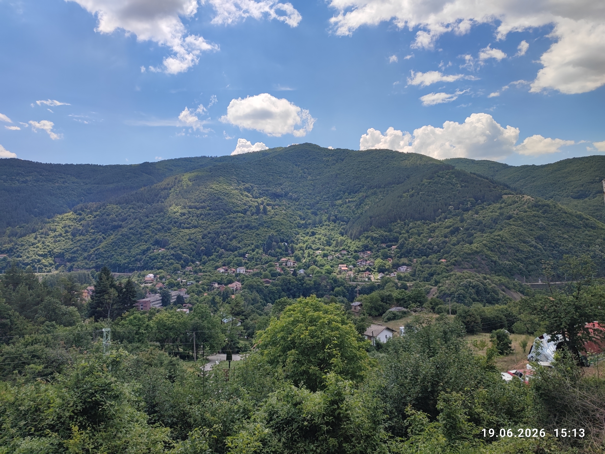 This media file captures a scenic view of a village nestled within a lush, green mountainous landscape under a bright, partly cloudy sky. The scene is dominated by the verdant slopes of the mountains, densely covered with trees. A small village is visible, with clusters of houses scattered among the trees on the hillsides. In the foreground, there is a dense canopy of green foliage, with the tops of trees and bushes extending towards the viewer. Power lines and a green metal pole are also visible in the mid-ground, suggesting the presence of infrastructure. On the far right, a dilapidated structure with a red, corrugated metal roof is partially visible, with some vehicles, including what appears to be a white car and a red car, parked nearby. The overall impression is one of a peaceful, rural setting in a mountainous region. The time stamp "19.06.2026 15:13" indicates the photograph was taken on June 19, 2026, at 3:13 PM, suggesting it is mid-afternoon on a sunny day, consistent with the bright lighting and scattered clouds. The location context places this scene in Tompsan, Bulgaria. No people are visible in the image.