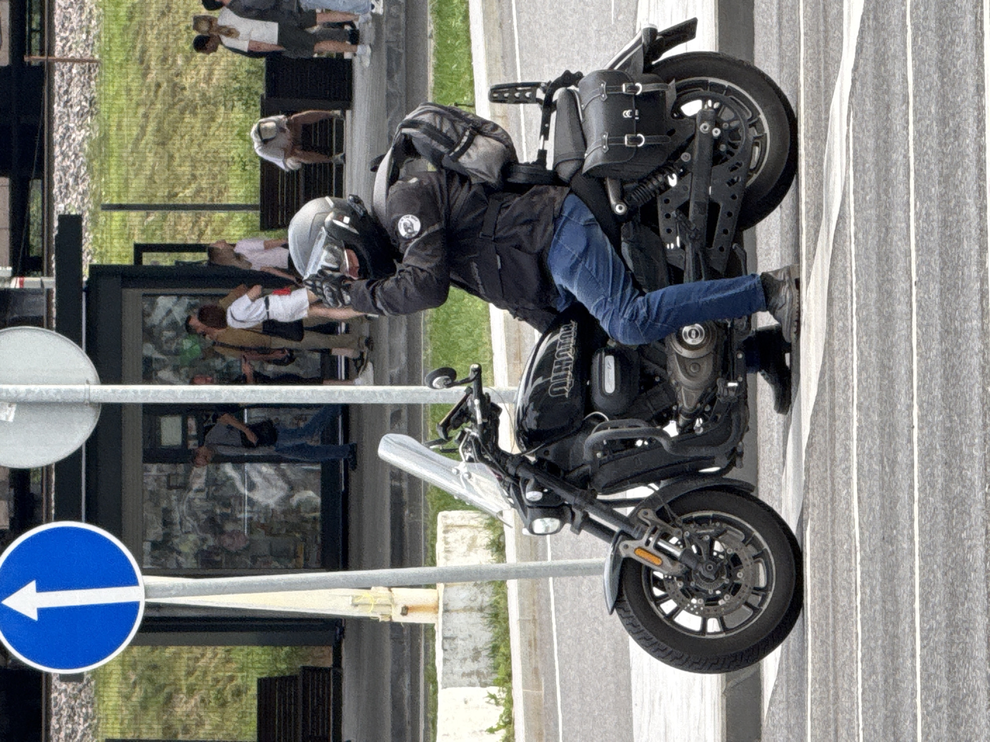 The media file shows a motorcycle with a rider in motion, captured from a slightly elevated side angle. The rider, wearing a black jacket, blue jeans, a helmet, and gloves, is leaning forward over the handlebars. A backpack is secured behind the rider. The motorcycle is dark-colored and appears to be a cruiser style, with saddlebags mounted on the rear. The scene takes place on a paved surface, possibly a street or sidewalk, with a grey, ribbed wall or building in the background. There are some metal railings and a circular blue sign with a white arrow pointing left visible in the foreground, suggesting a traffic or pedestrian area. In the background, through a window or glass partition, several people can be seen in what appears to be an indoor setting, some standing and others possibly sitting. There are also glimpses of greenery and a rocky texture, potentially indicating an urban environment with some natural elements or landscaping. The lighting suggests it is daytime. The overall impression is of a candid shot, possibly capturing a moment during a motorcycle ride or a pause in activity. Text visible on the motorcycle's engine area reads "HARLEY DAVIDSON".