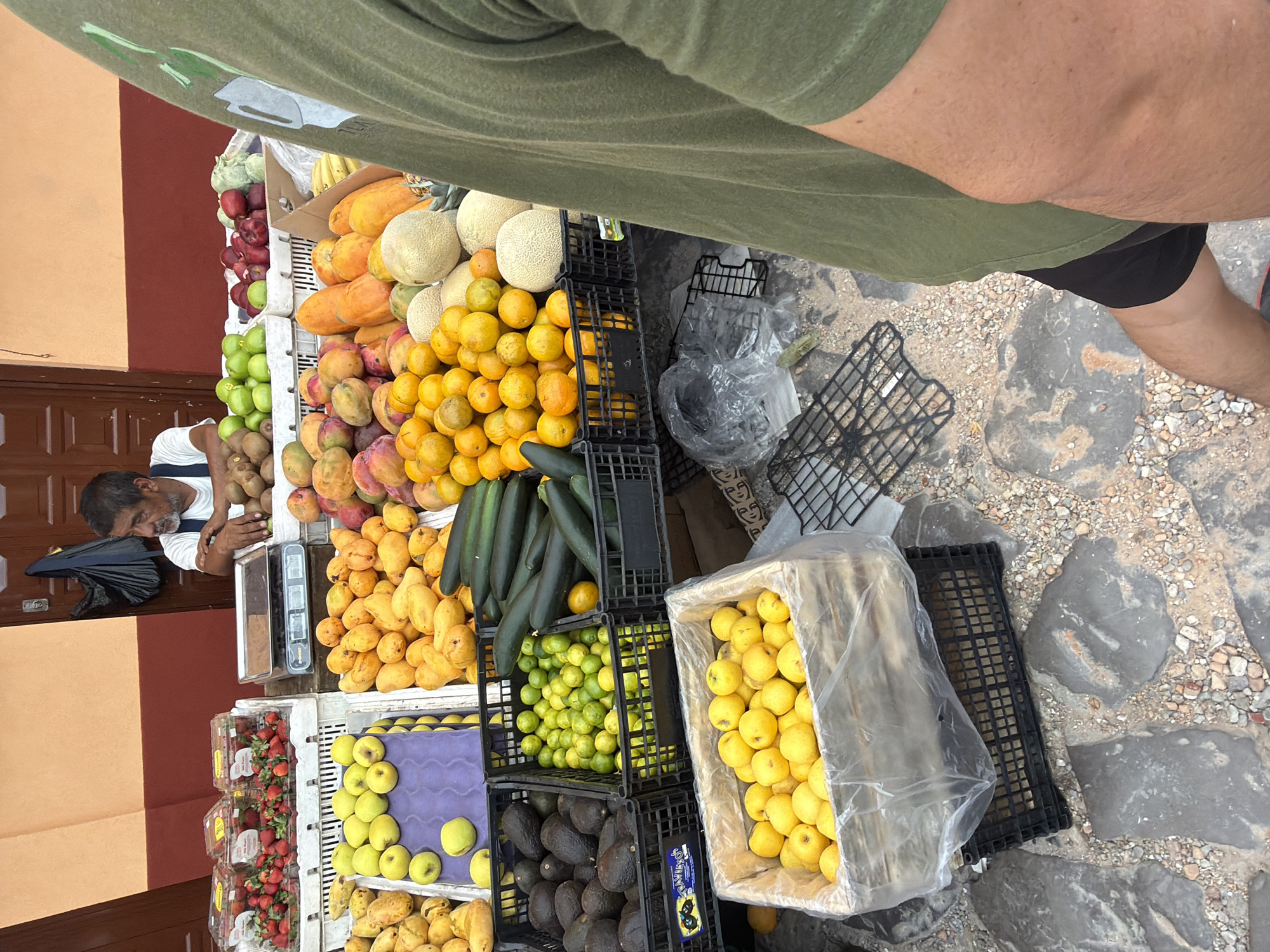 The image shows a fruit and vegetable stand in Cuetzalan, Mexico. An older man, presumably the vendor, is resting at the far end of the stand. The stand is filled with various fruits and vegetables, including strawberries, apples, apricots, mangoes, kiwi, oranges, cantaloupe, papaya, zucchini, limes, lemons, and avocados. A person wearing a green shirt and black shorts is standing at the stand, likely a customer. The stand is located on a stone pavement.