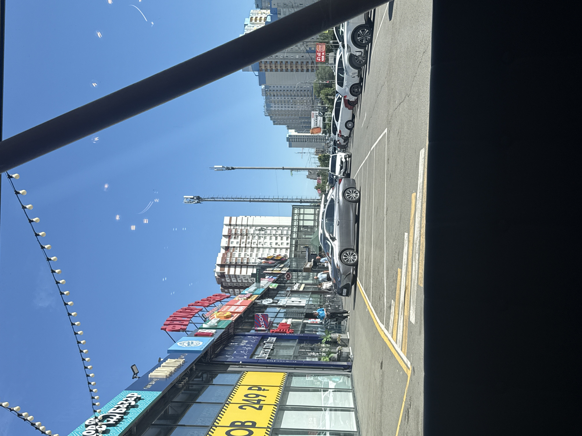 This image depicts a view of a commercial strip with a parking lot in front, observed from inside a vehicle. The scene is set in Orenburg, Russia, specifically in the area of the shopping center "Grad", located at 154 Vostochnaya Street. The architecture suggests a modern commercial development, with multi-story buildings lining the street and a clear blue sky overhead.
In the foreground, a gray sedan is parked in a designated spot, facing away from the viewer. The parking lot is paved and marked with white and yellow lines. Beyond the parking lot, a row of shops with large glass windows and various signs is visible. Some signs are in Russian, advertising goods and services such as "OB 249 P", "ЦВЕТЫ ДЛЯ ТЕБЯ" (Flowers for you), and "OZON". People are seen walking along the sidewalk in front of the shops, suggesting active commercial activity.
Further in the background, taller residential buildings dominate the skyline, characteristic of urban development. Two cellular towers are also visible, indicating communication infrastructure in the area. Reflections on the glass of the vehicle from which the photo is taken are noticeable, including streaks of light and faint outlines of overhead lights. The overall impression is a typical sunny day in a commercial district of a Russian city.