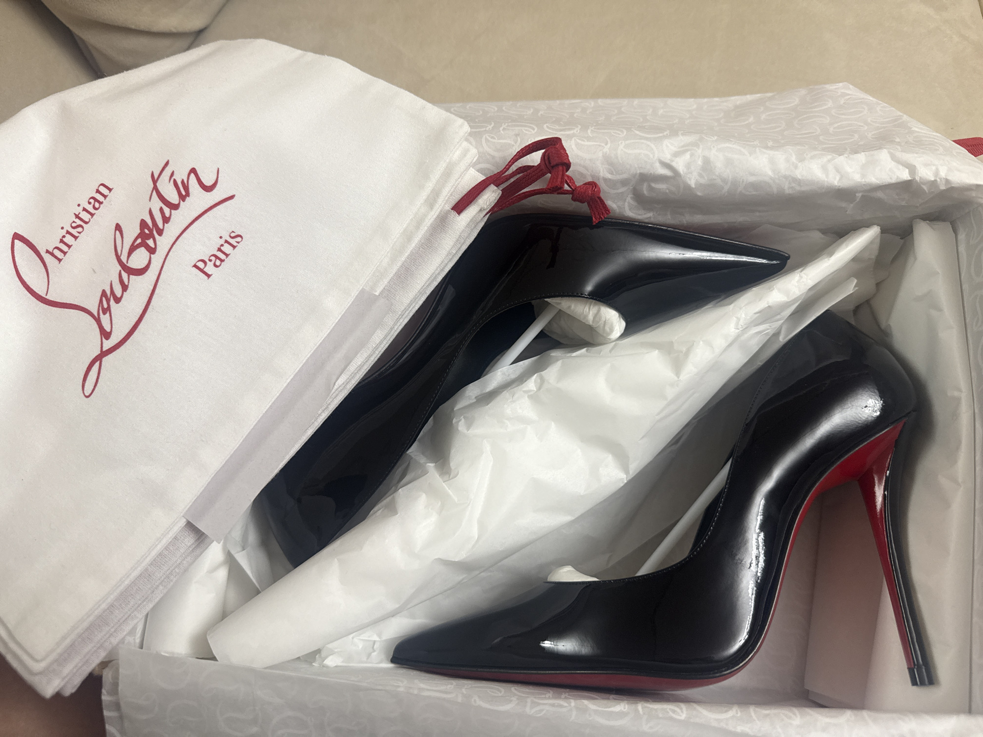 The image shows a pair of black patent leather stiletto heels with red soles and red heel tips, likely Christian Louboutin brand. They are nestled in a white shoe box with white tissue paper. A white dust bag with the "Christian Louboutin Paris" logo in red lettering is partially visible next to the shoes. The red drawstrings of the dust bag are visible. The box has a white pattern consisting of small white symbols. The environment appears to be indoors, possibly a residence. There is a tan colored item in the background. The location in the city of Hallandale Beach, United States of America cannot be determined from this image.
