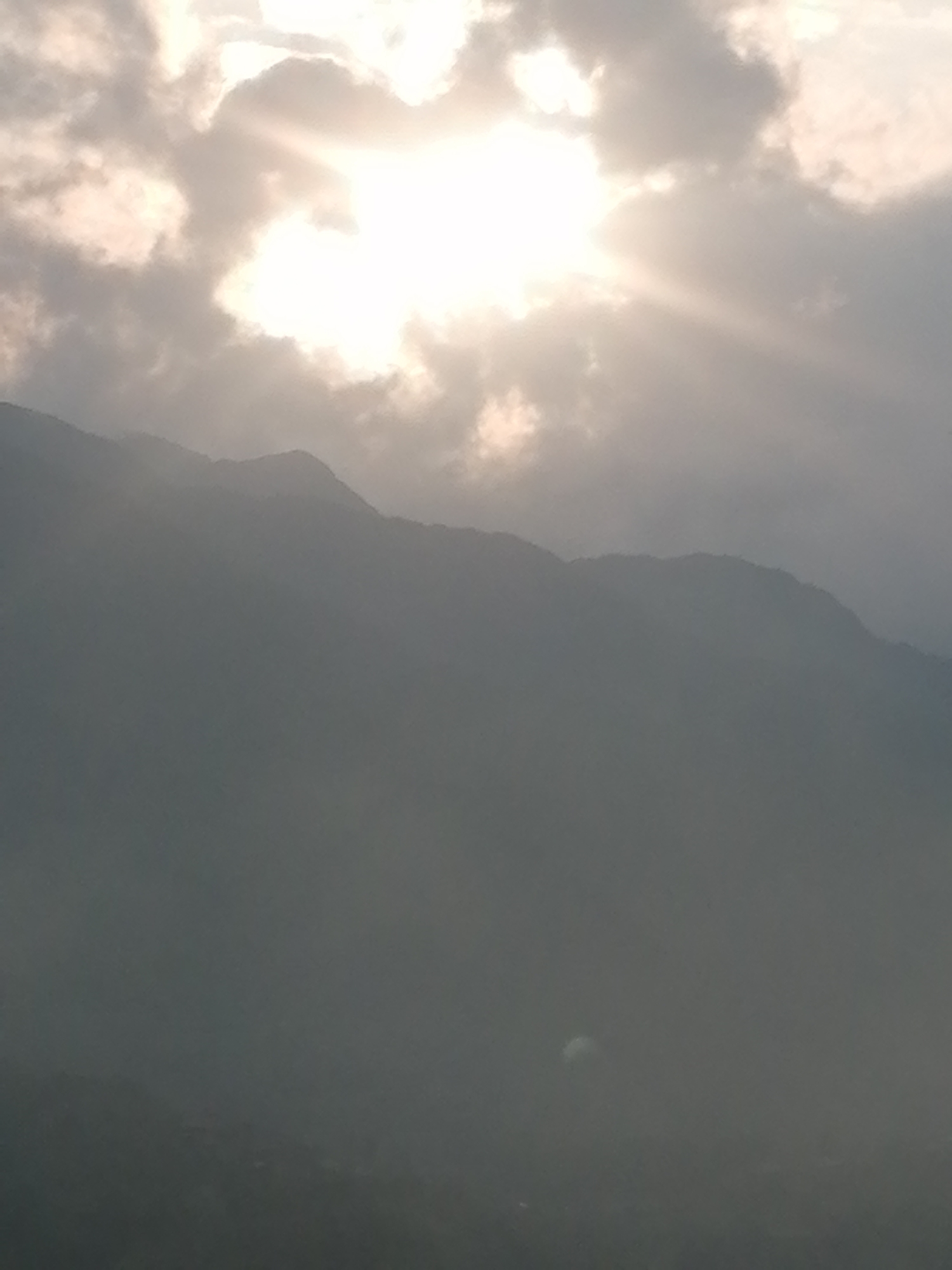 The image shows a landscape in the Sa Pa Ward, Vietnam. A mountain range is partially obscured by mist. Above the mountains, clouds are visible, with the sun shining brightly through a gap in the cloud cover. Rays of light extend from the sun. The lower portion of the image is dark, possibly due to shadows or fog.