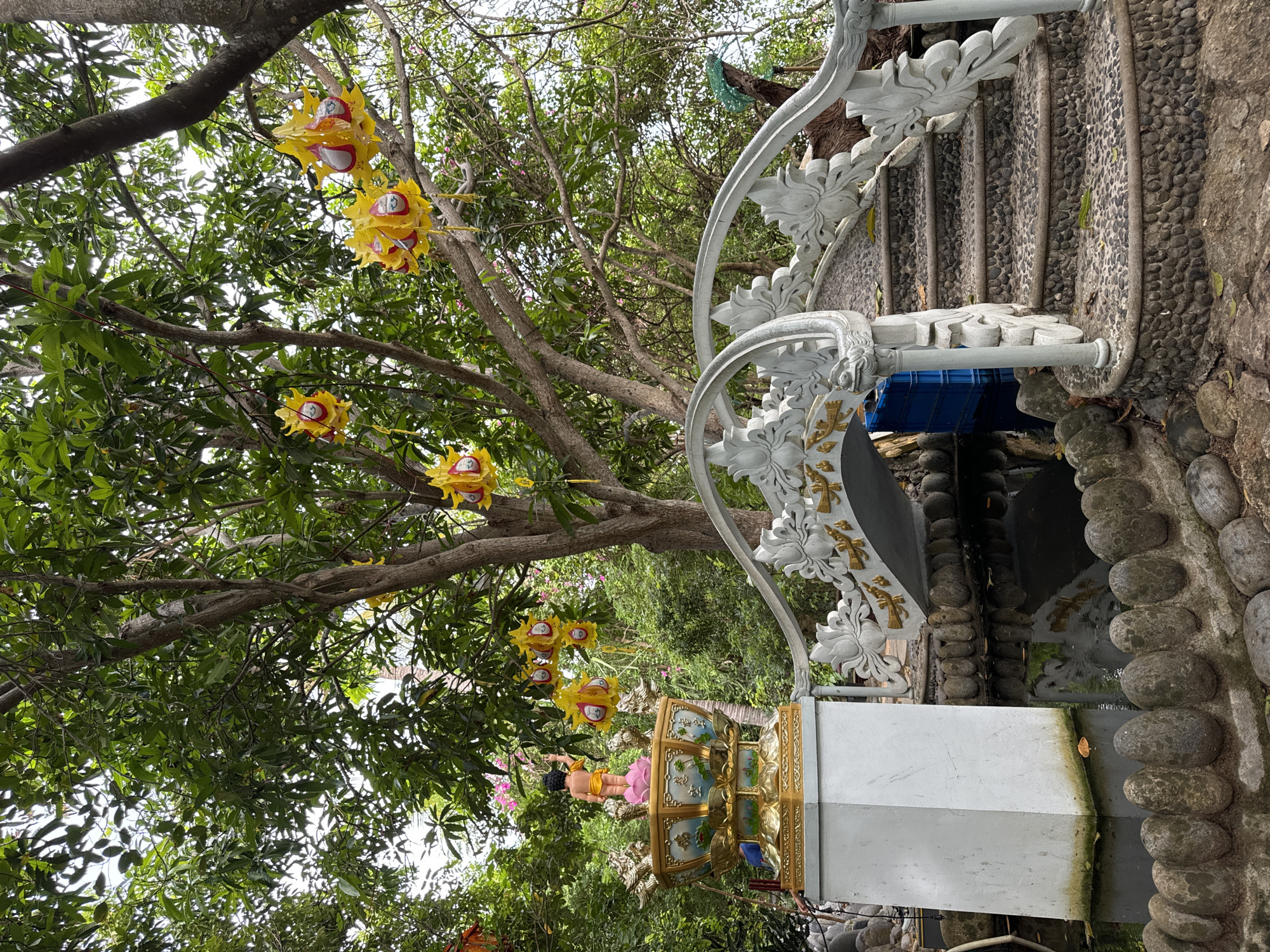 The image depicts an outdoor scene, likely a garden or temple area, with lush green foliage and trees. Hanging from the branches are several bright yellow, lantern-like decorations. In the foreground, a white, ornate bridge with detailed carvings, possibly depicting lotus flowers and dragon heads, spans over a calm body of water. Steps paved with small, dark pebbles lead up to the bridge on the right side. To the left of the bridge, a white pedestal supports a golden, ornate basin topped with a statue of a young child, likely Buddha, standing on a pink lotus. The background is filled with dense greenery, suggesting a tropical environment. The overall impression is one of tranquility and cultural significance, potentially a religious or spiritual site. Given the style of the decorations and the statue, the location could be a Buddhist temple or shrine in Nha Trang, Vietnam.