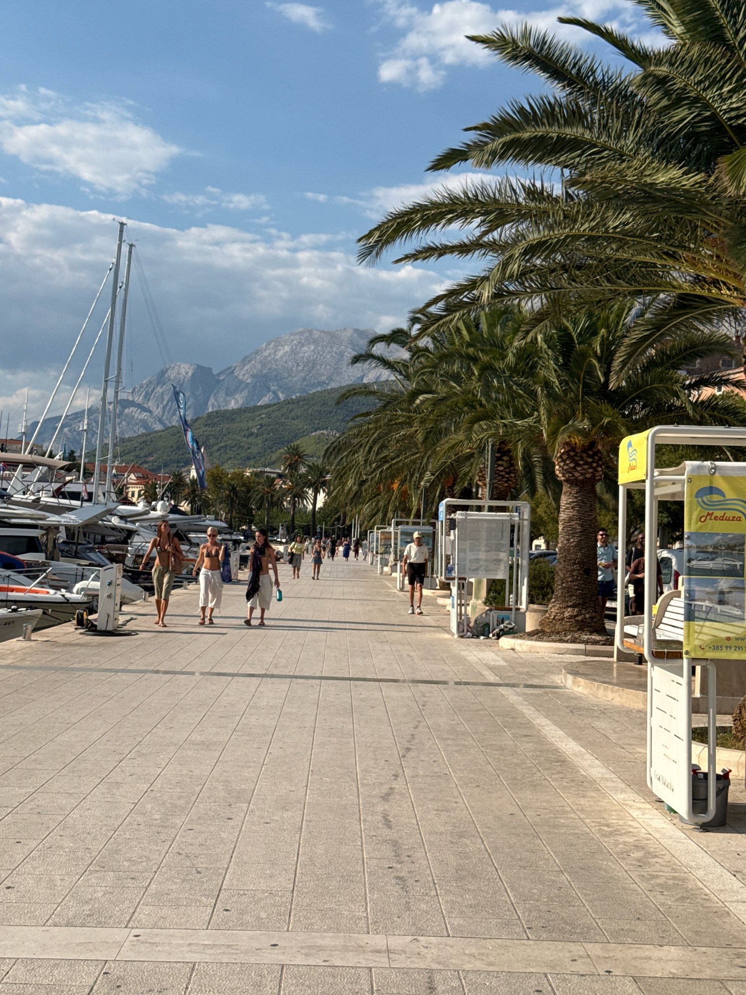 The image depicts a promenade in Makarska, Croatia, with a harbor on the left and a row of palm trees on the right. Several yachts are docked in the harbor. In the foreground, three women are walking away from the camera on the paved promenade. Behind them, other people are walking, and some are standing. In the background, mountains rise above the city. The sky is blue with some clouds. The lighting suggests it is daytime, likely late afternoon or early evening, given the long shadows cast by the palm trees and the warm light. Several information stands or kiosks are present along the promenade, one of which displays the word "Meduza".