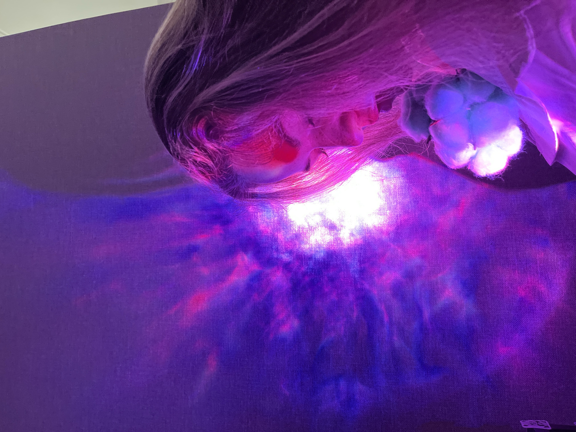 A person with light hair is positioned in front of a purple background. A bright, diffuse light source illuminates the person's face and the surrounding area, creating colorful patterns of pink, blue, and purple on the background. A cotton-like object is located near the person's face. There is no information available about the location in the city of Chaikovsky, Russia.
