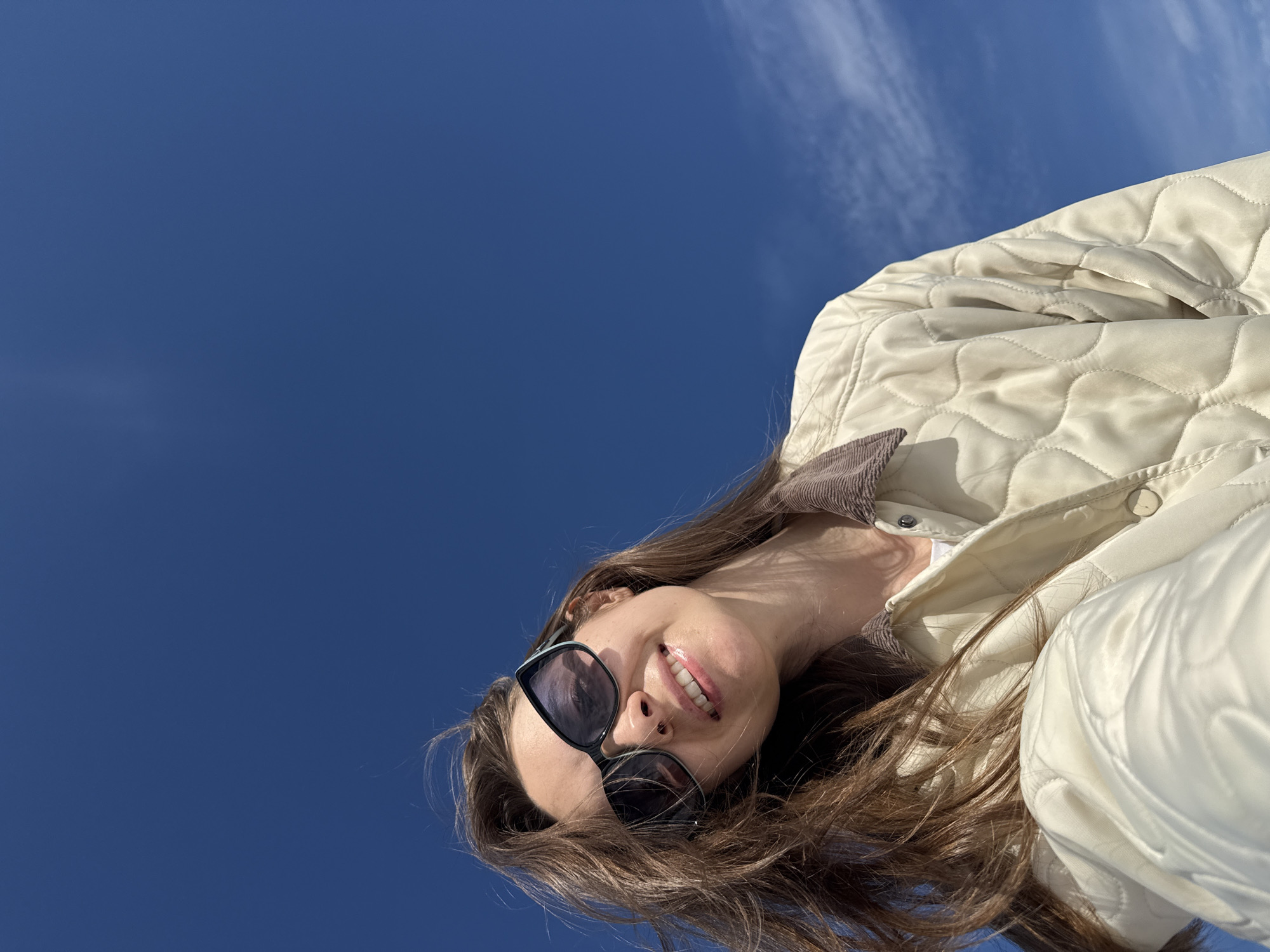 The image displays an adult female in a low-angle, upward-looking perspective, presumably taken as a selfie. She is located in Chaikovsky, Russia. Her long, dark brown hair is styled back and appears slightly windblown. She wears dark-lensed, angular sunglasses, obscuring her eyes. Her mouth is open in a smile, revealing her teeth. Her attire consists of a light cream or beige quilted jacket with a contrasting dark brown corduroy collar, worn over a white garment visible at the neckline. The background is a clear, deep blue sky with a few faint, wispy white clouds visible primarily in the upper and lower right quadrants. The lighting is bright, indicating daylight conditions.