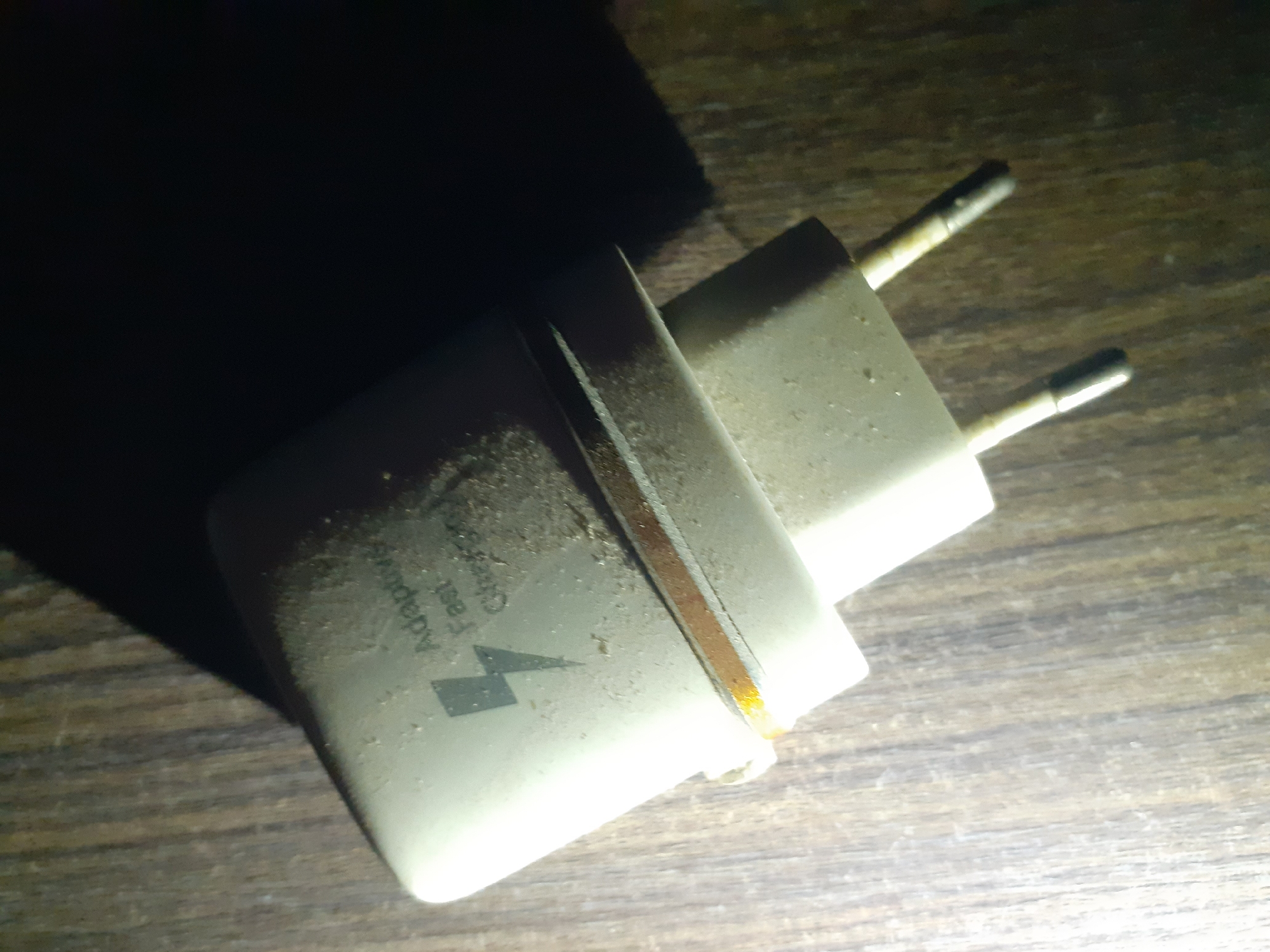 The image shows a close-up, high-angle shot of a white Samsung Adaptive Fast Charger, covered in dust, lying on a wooden surface. The charger is angled, with the plug prongs facing downwards and to the right. A distinctive lightning bolt symbol and the words "Adaptive Fast Charging" are visible on the side of the charger, though partially obscured by dust. The wooden surface appears to be a table or floor, with visible grain patterns. The lighting in the image is dim, with a strong light source casting a shadow from the charger and obscuring part of the background. The scene is static and appears to be indoors, in a setting that is not well-maintained due to the presence of dust. There is no indication of people, actions, or interactions. The location in Jalingo, Nigeria is not discernible from the image itself.