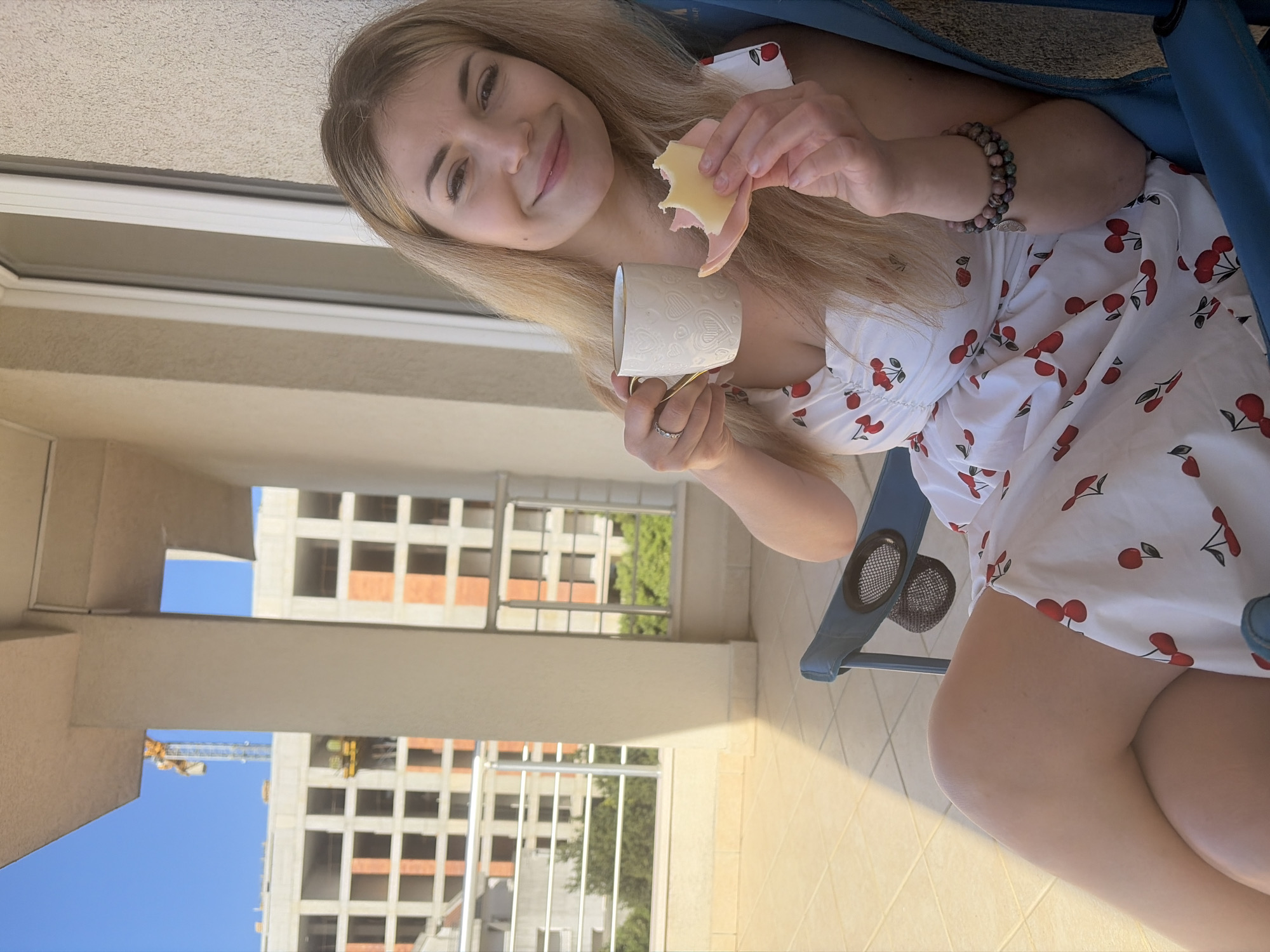 A young woman sits in a blue folding chair on a balcony, holding a white heart-patterned mug in her left hand and a ham and cheese sandwich in her right. She is fair-skinned with long blonde hair, wearing a white dress with a cherry pattern. She looks at the camera and smiles.
The balcony overlooks a construction site. Unfinished buildings with exposed brick are visible across the frame. A yellow crane stands among the construction. The scene takes place in Budva, Montenegro, based on the building style and environment.
