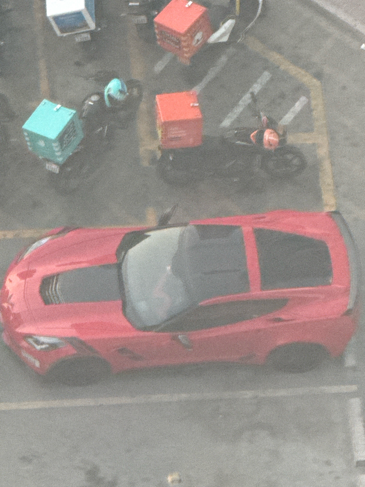 The image, taken from an elevated perspective, shows a red sports car parked on asphalt pavement marked with white and yellow lines, possibly indicating parking spots. The car is oriented facing away from the viewer, with its front end on the left and rear on the right. Several delivery scooters with insulated cargo boxes are visible in the background. One scooter, positioned behind and to the left of the sports car, has a teal-colored box. Another scooter, further back and to the right, has a red cargo box. A person wearing a helmet is seated on this scooter. To the left of the red sports car, another scooter with a teal cargo box is parked. Further to the left and behind, a white and blue modular structure is visible. The scene appears to be an outdoor urban setting. The atmospheric conditions are hazy or dusty, affecting the clarity of the image.
