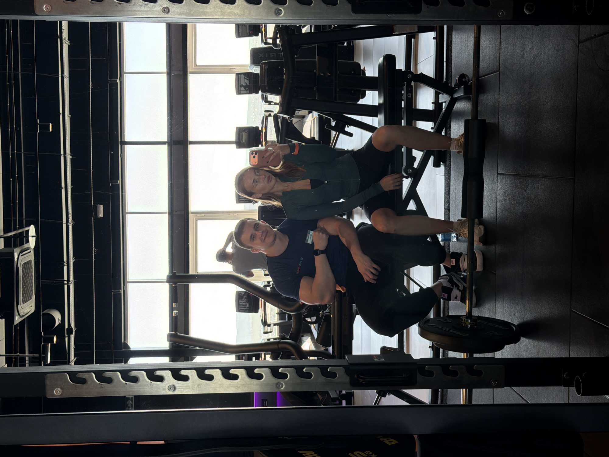 This media file is an image, rotated 90 degrees clockwise, depicting a mirror selfie taken by a woman with a man beside her in a gym setting.
**Subjects:**
* **People:** A young woman with long, light brown hair, wearing a dark green athletic top and black shorts, is holding a smartphone to take the picture. Beside her, a young man with short, dark hair is wearing a dark blue t-shirt and dark athletic pants.
* **Objects:** The woman is holding a light-colored smartphone. Various gym equipment is visible, including a squat rack with a barbell and a weight plate on the floor, benches, and other exercise machines in the background.
* **Scene/Setting:** The scene is a modern gym, characterized by the dark-colored walls, black floor mats, and extensive fitness equipment. Large windows in the background allow bright daylight into the space.
**Activity or event taking place:**
The woman is taking a mirror selfie, capturing herself and the man in what appears to be a casual pose during or after a workout.
**Notable details:**
The bright light streaming through the large windows suggests it is daytime. Both the woman and the man are smiling, indicating a happy and relaxed mood. The gym appears well-lit and spacious. The overall impression is one of a positive and active lifestyle.
**Visible text:**
On the man's t-shirt, the words "SIBIRIAN Health" are visible. On some gym equipment in the background, part of a brand name, possibly "ATRIX" or "XTR," can be seen.
