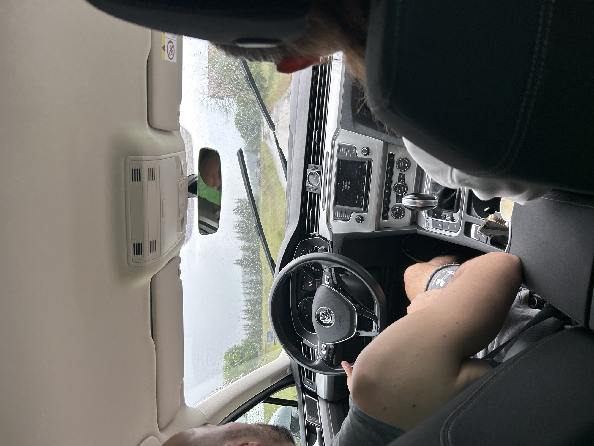 The image shows the interior of a car, likely a Volkswagen, viewed from the back seat looking forward. The scene is dominated by the car's interior, including the dashboard, steering wheel, and parts of the seats. Two individuals are visible. One person, likely the driver, is in the foreground, with their arm resting on the center console and their hand on the steering wheel. Another person is visible in the rearview mirror, reflected from what appears to be the passenger seat.
The car is on a road, as indicated by the windshield view showing trees and a cloudy sky. The weather appears to be overcast and possibly rainy, as streaks are visible on the windshield. The location is identified as Cherna Mesta, Bulgaria.
Notable details include the car's infotainment system displaying song information ("Procesal Jam," "High Ride," "High Ride"). The dashboard shows a speed of 55. A warning label with a "no entry" symbol is visible on the overhead console. The lighting suggests it is daytime, but the cloudy weather creates a diffused light. The overall mood is relaxed, with the individuals seemingly on a journey.