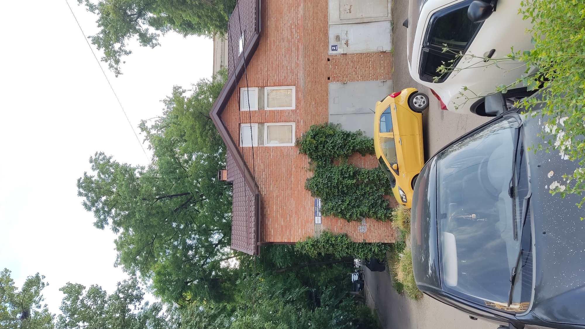 The image shows a residential area in Krasnodar, Russia. A two-story brick building with a brown roof is visible, featuring multiple windows and two garage doors. Ivy covers parts of the brick facade. A yellow hatchback car is parked in front of the building, with its front end facing left. In the foreground, a dark-colored car is parked, its hood and windshield partially visible. Another white car is parked to the right of the dark-colored car. Trees and greenery surround the building and line the adjacent street. The sky is overcast. A street sign with Cyrillic text, "107/7 СТАВРОПОЛЬСКАЯ", is affixed to the brick wall.