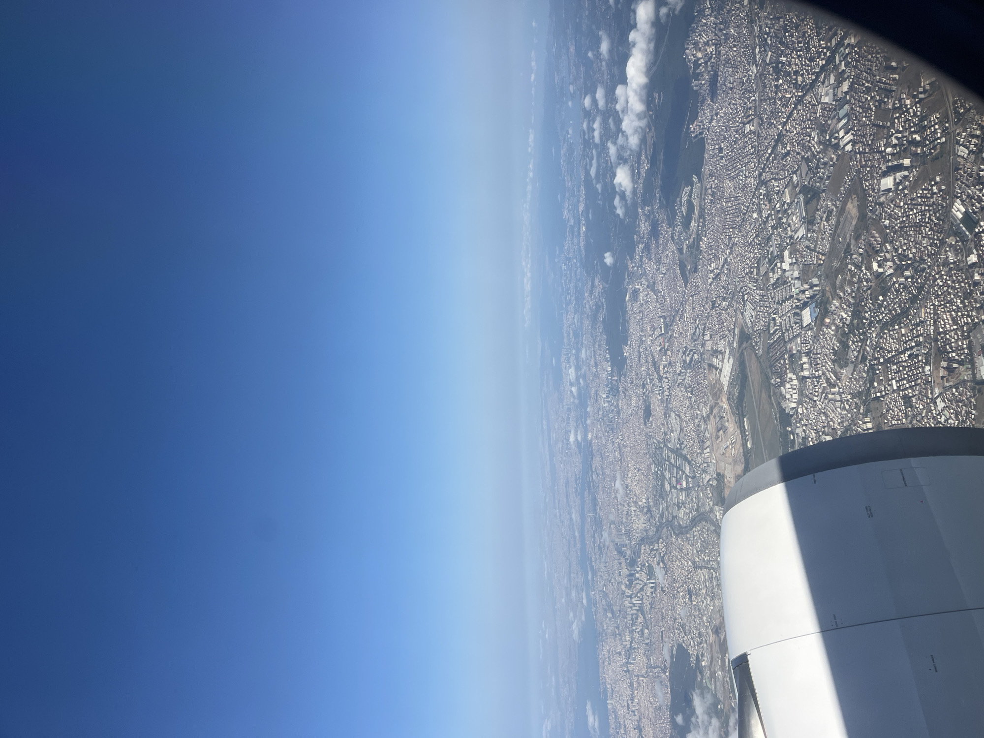 An aerial view captures a dense urban landscape under a clear blue sky, with the engine nacelle of an airplane visible in the foreground, partially obscuring the view. The city stretches out to the horizon, characterized by a vast expanse of buildings with a mix of red-tiled roofs and grey structures. Roads and infrastructure are visible, weaving through the urban sprawl. Scattered clouds dot the sky and cast shadows on the city below. The scene appears to be a daytime aerial shot taken from an aircraft in flight.