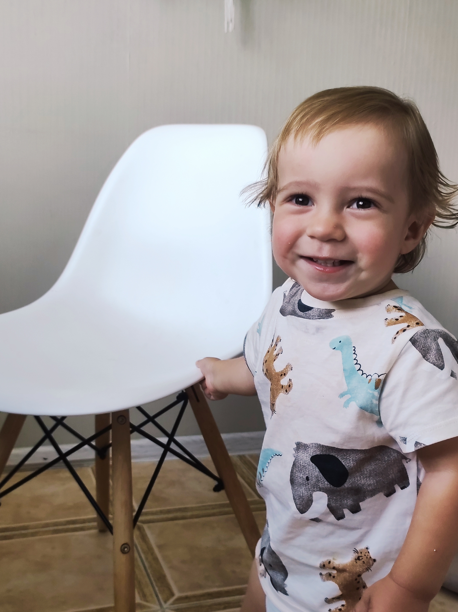A young child, appearing to be between 1 and 3 years old, with blonde hair and fair skin, smiles at the camera. The child is wearing a light-colored t-shirt with a repeating pattern of cartoon animals, including elephants, dinosaurs, and what appear to be felines. The child's left hand is visible, resting on the edge of a white molded plastic chair with wooden legs. The chair has a black criss-cross support structure between the legs. The background consists of a plain light-colored wall and a tiled floor with a geometric pattern. The scene is indoors, possibly in a residential setting. The location of Chaikovsky, Russia is not discernible from the image.