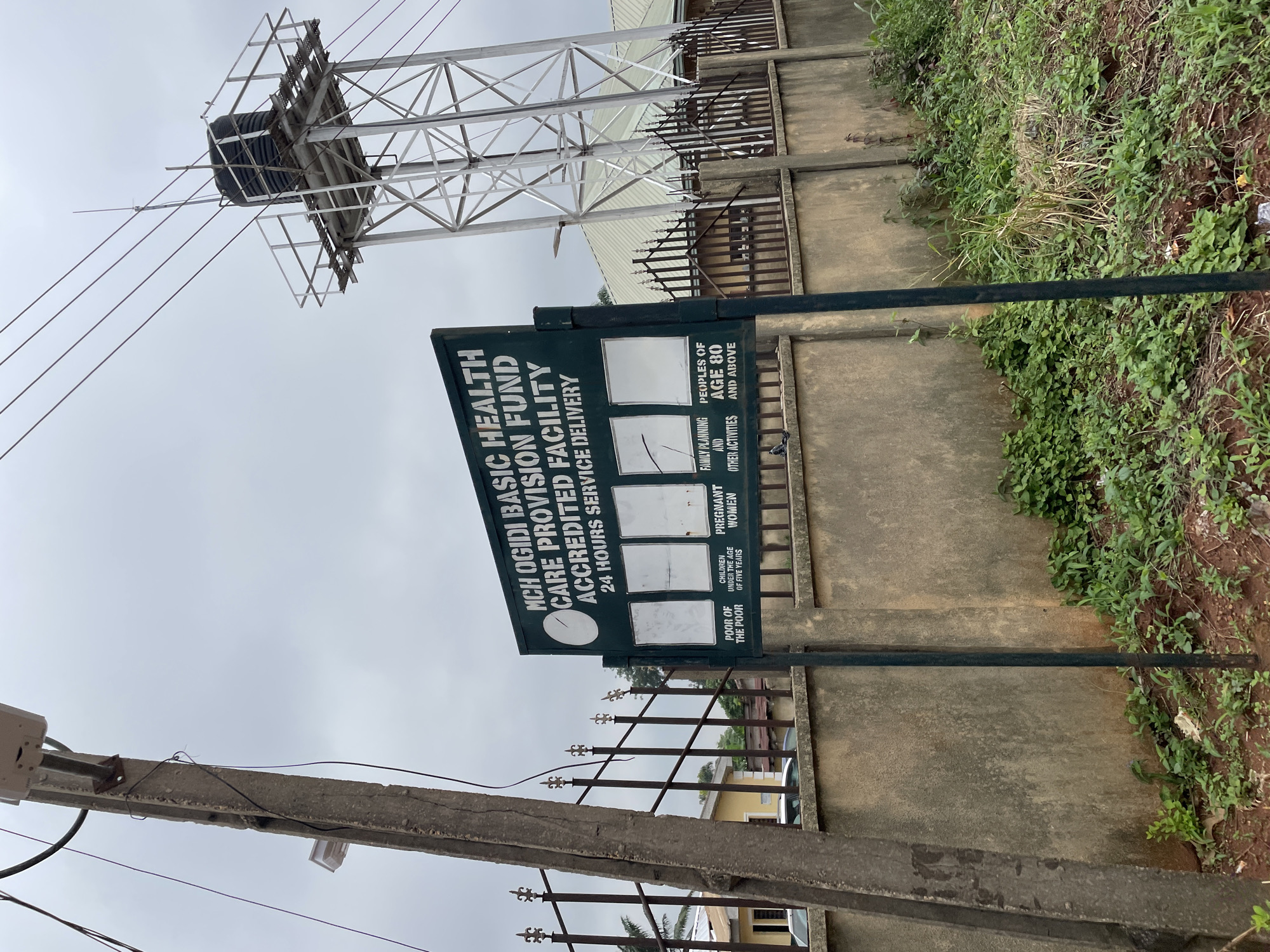 This image depicts an outdoor scene in Ogidi, Nigeria, featuring a sign for the MCH Ogidi Basic Health Care Provision Fund Accredited Facility. The sign is dark green with white lettering and indicates "24 HOURS SERVICE DELIVERY." Below the main text, there are sections for various services, including "POOR OF THE POOR," "CHILDREN UNDER THE AGE OF FIVE YEARS," "PREGNANT WOMEN," "FAMILY PLANNING AND OTHER ACTIVITIES," and "PEOPLES OF AGE 80 AND ABOVE." Several blank panels are visible, suggesting they are intended for further information or updates.
In the background, a tall metal water tower with a black tank is prominent. A building with a light-colored, sloped roof and a dark metal fence is situated behind the sign. To the left of the sign, a concrete utility pole with electrical wires and some attached fixtures is visible. The area around the sign and the building appears to be overgrown with vegetation and grass, with a concrete wall running along the foreground. The sky is overcast, suggesting a cloudy day. No people are visible in the frame.