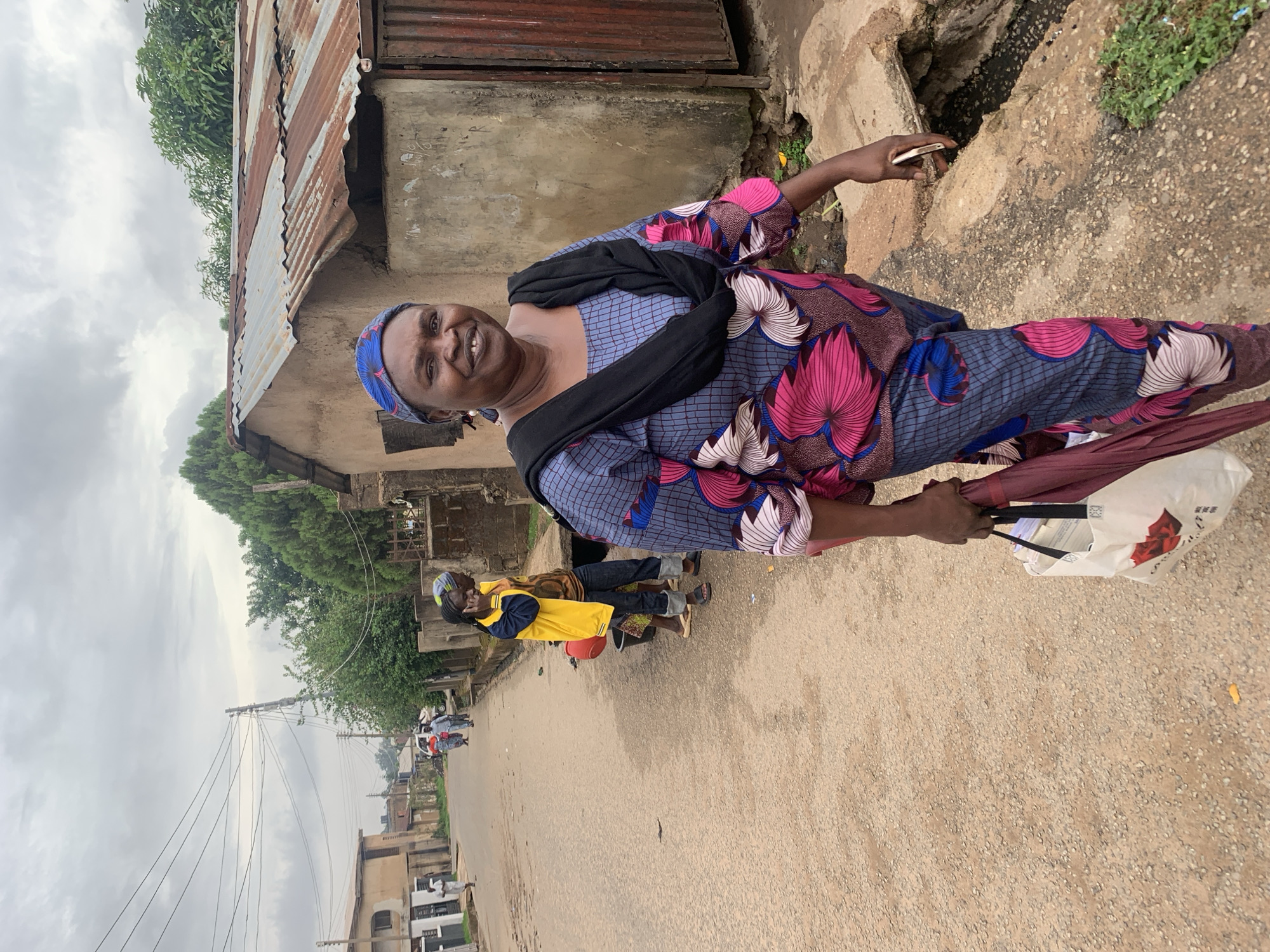 A woman stands on a street in Jos, Nigeria. She wears a blue, pink, and white patterned dress, a black headscarf, and a black shawl. She smiles while holding a cell phone in her right hand and a bag and umbrella in her left hand. Behind her is a building with a rusted corrugated metal roof and an exposed concrete foundation. Another person is visible further down the street wearing a yellow jacket. Power lines are above the street.