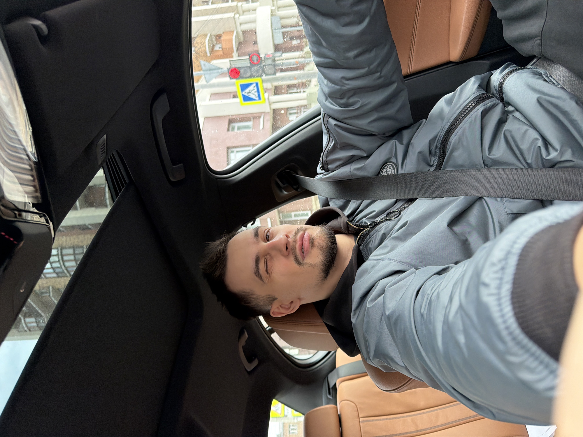 A man is in a car, wearing a gray jacket and a seatbelt. The interior of the car features brown leather seats and a sunroof. Through the sunroof, a street scene is visible, including a red traffic light and a blue and yellow crosswalk sign. The location is identifiable as Krasnoyarsk, Russia, by the distinct style of traffic signage. The man has one eye closed and the other eye open.