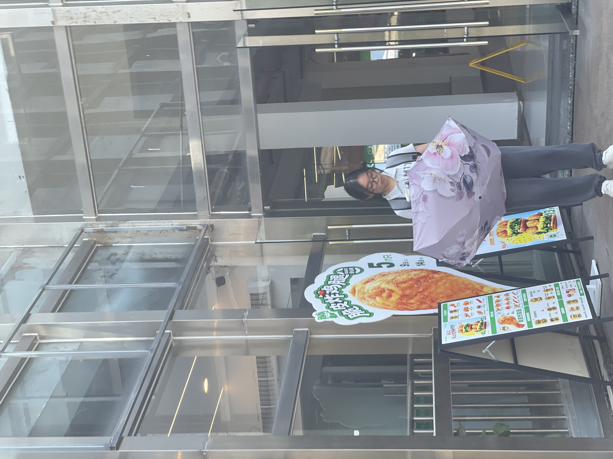A young woman stands in front of a storefront in the High-tech Zone of Chongqing, China. She wears glasses, a striped shirt, and gray pants, and holds a folded umbrella with a floral design. Behind her, signs display food items, likely advertising the restaurant or store. The building facade is predominantly glass and metal.