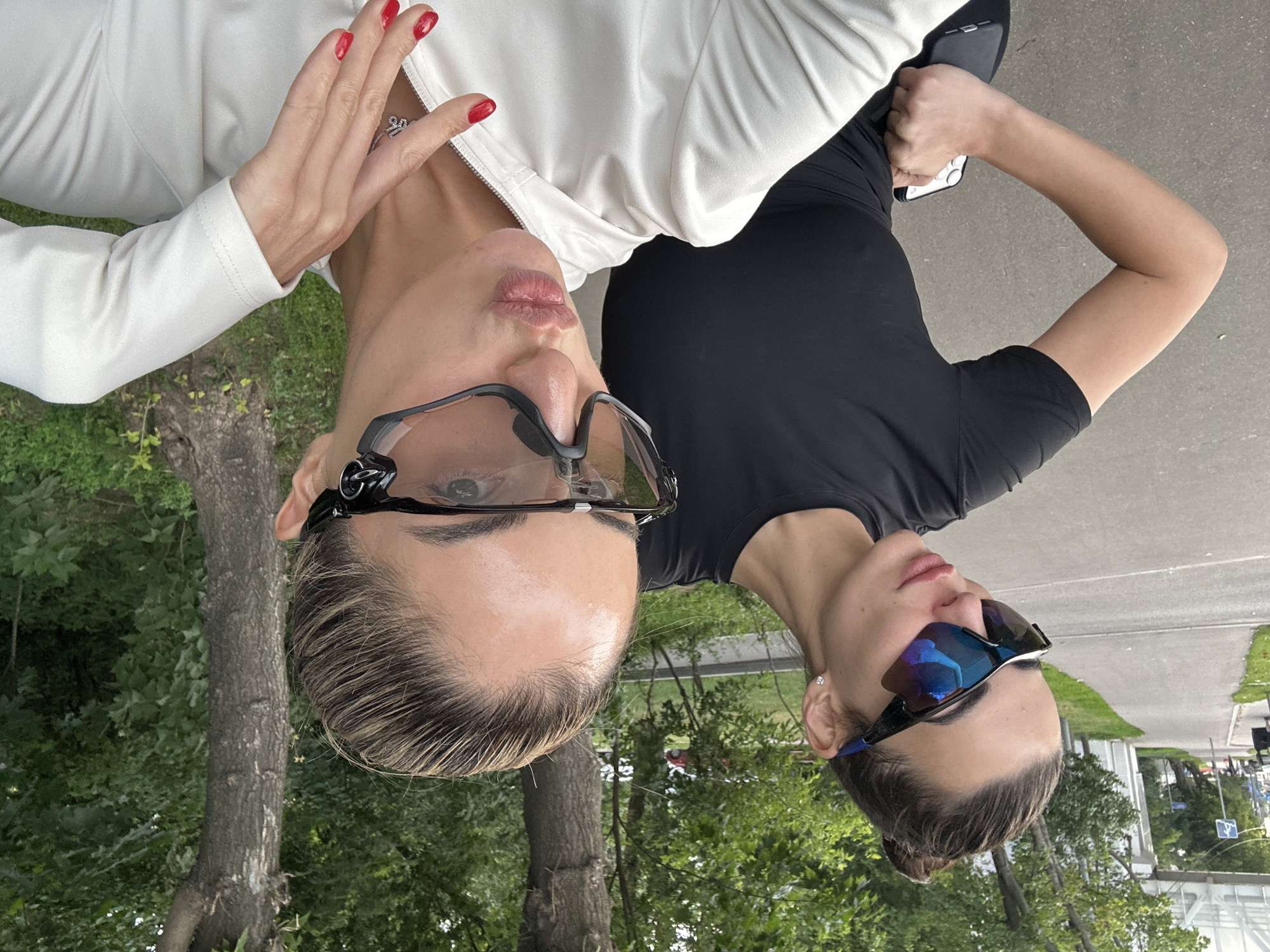 The media file shows a close-up selfie of two women outdoors, with their faces angled towards the camera and the image rotated 90 degrees counterclockwise. One woman is wearing a white jacket and has bright red painted nails, with her hand touching her neck. She is also wearing dark sunglasses. The second woman, partially visible, is wearing a black t-shirt and also has dark sunglasses. Both women appear to be wearing makeup.
The setting is outdoors, with lush green trees and foliage in the background. Part of a paved walkway is also visible, suggesting a park or public area. The lighting indicates it is daytime, with bright, natural light.
The women are looking at the camera, and the woman in the white jacket is pouting her lips, suggesting a playful or posed expression. There are no visible signs of text within the image. The overall impression is a casual, outdoor photo session between two friends. The location context is Moscow, Russia.