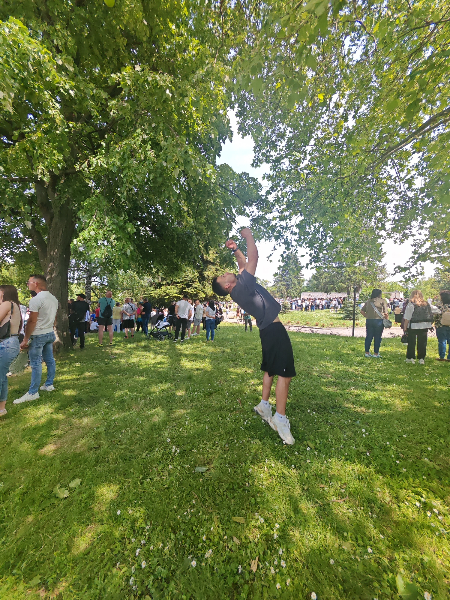 The image shows a man in a black t-shirt and black shorts performing a backbend in a grassy area. He is wearing white sneakers and his face is tilted upwards. Several people are standing around in the background, and trees provide shade. The location is in the city of Shumen, Bulgaria. The grass has small white flowers scattered throughout. The people in the background appear to be gathered in small groups. Some are standing near trees. A stroller is visible amidst the crowd.