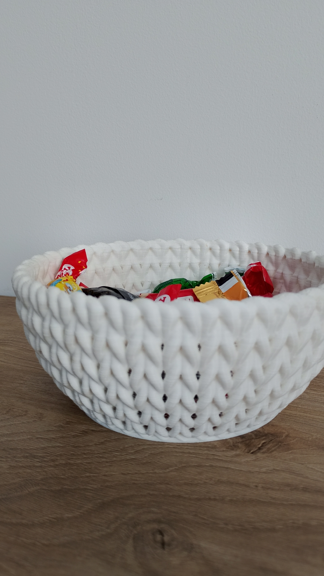 A white knitted basket filled with individually wrapped candies is placed on a wooden surface. The basket has a textured pattern with small openings. The candies have colorful wrappers, including red, green, gold, and yellow. The background is a plain white wall. This scene is not located in the city of Smolyan, Bulgaria.