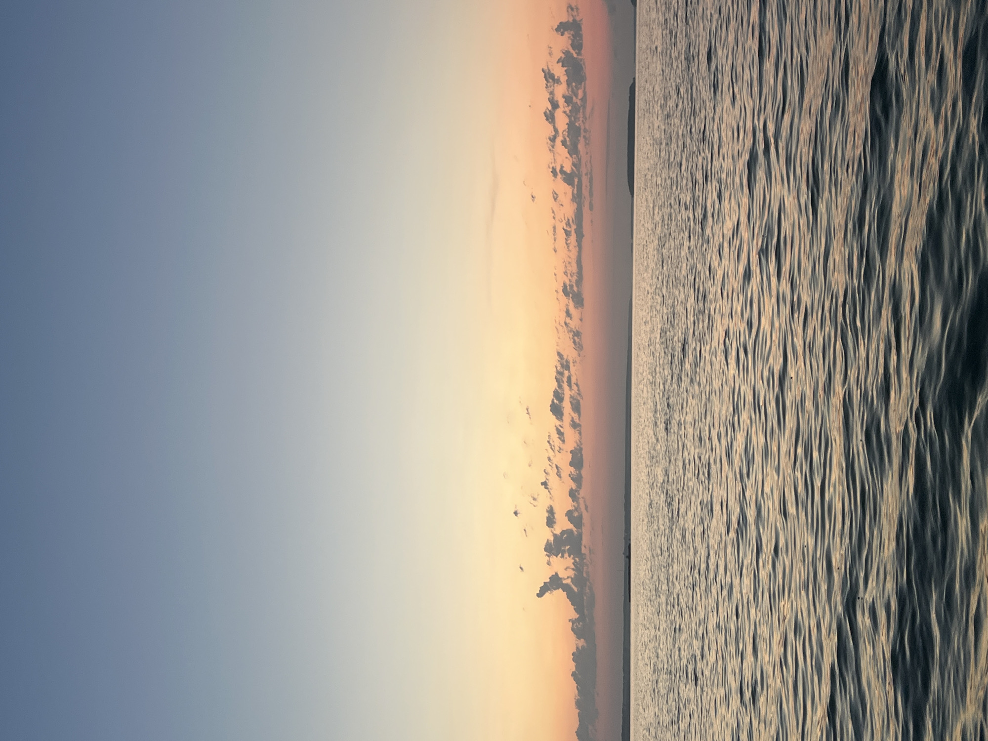 This is a landscape photograph taken at dusk. The image is oriented vertically. The left side of the image features a gradient of the sky, transitioning from a light blue at the top to a soft orange and pink near the horizon. Scattered, dark clouds are visible along the horizon line. The right side of the image depicts a body of water with gentle, rippling waves. The water reflects the colors of the sunset, giving it a warm, golden hue mixed with darker tones. The texture of the water's surface is detailed, showing the patterns of the waves. In the far distance, a faint silhouette of land is discernible along the horizon. The scene evokes a sense of calm and tranquility associated with a sunset over water. The time of day is clearly evening, indicated by the sunset colors. There is no visible text in the image. The location context provided is Novopichugovo, Russia.