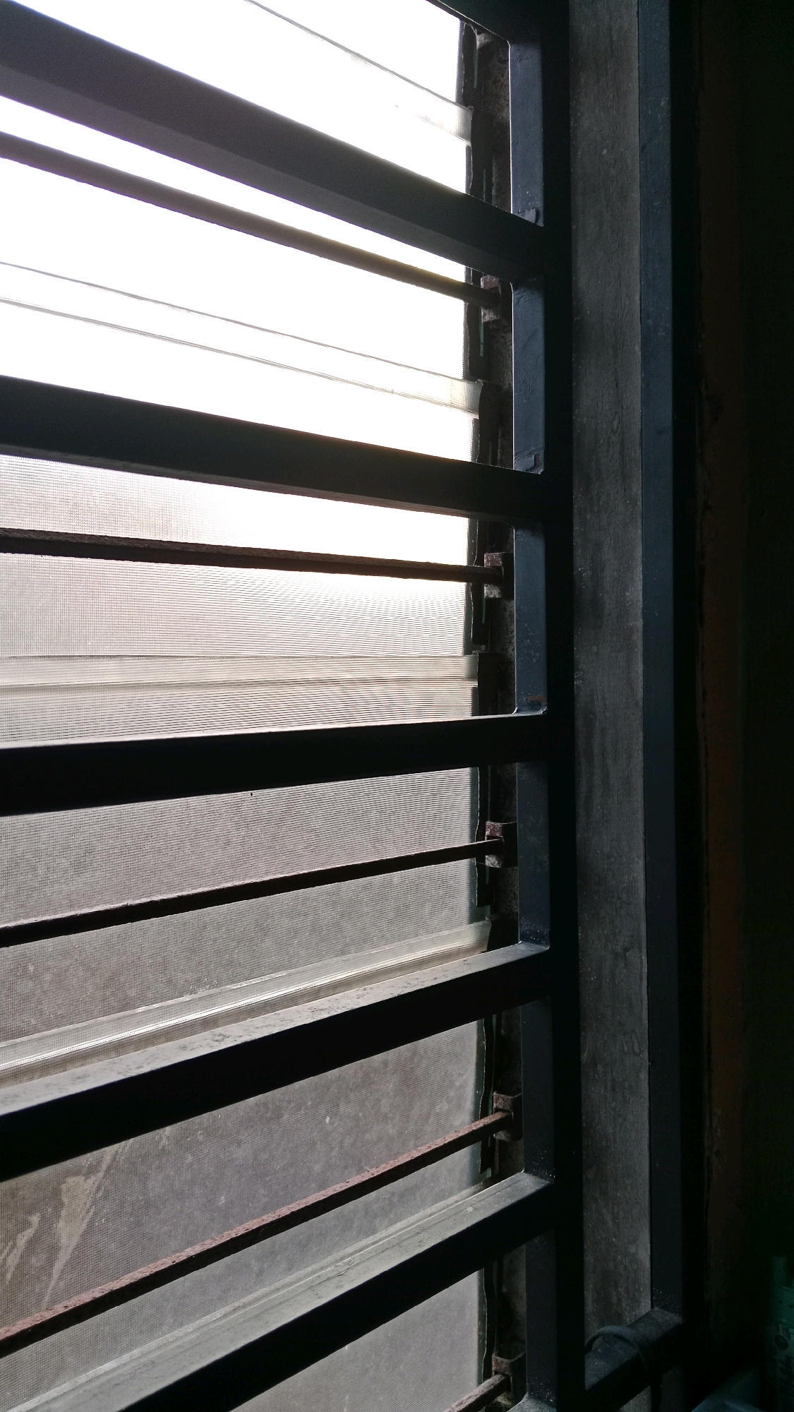 A close-up, interior shot shows a window with horizontal louvers covered by a screen. The louvers are dark and appear to be metal. The screen is translucent and dusty, allowing light to filter through. The window frame is also dark, with a vertical wooden beam visible on the right side. The overall scene is dimly lit, suggesting it is either early morning or late evening. The location is identified as Tanjung Pinang, Indonesia. There are no people or animals visible in the image. The focus is on the texture and light filtering through the window.