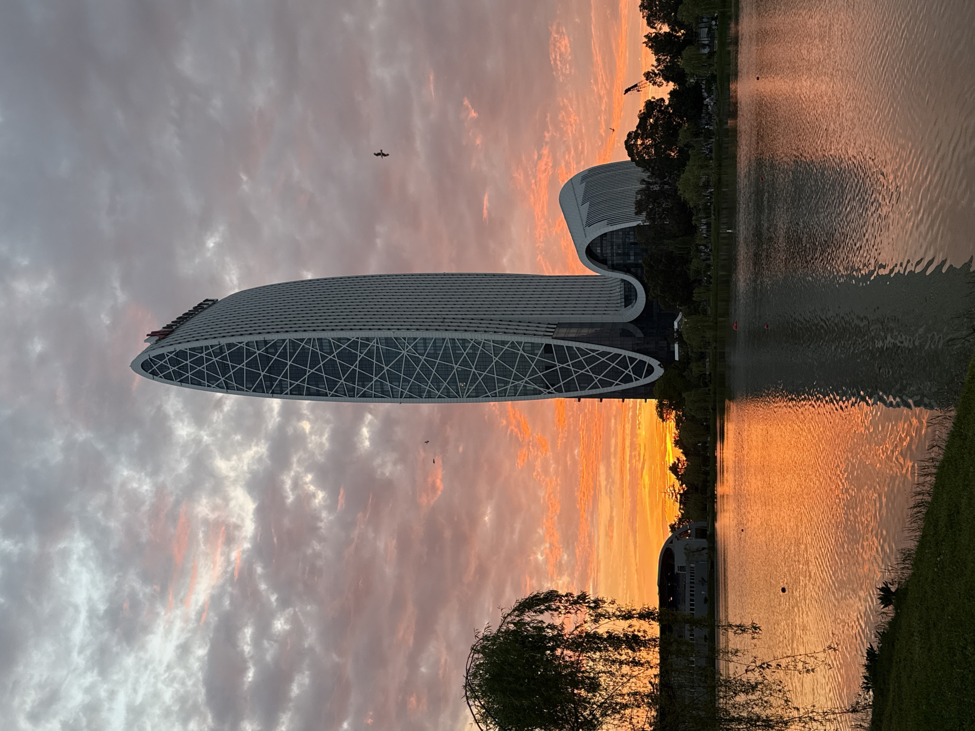 The image captures a scene in Batumi, Georgia, featuring the Radisson Blu Hotel. The building has a distinctive modern design with a curved shape and a patterned facade. A body of water, likely a lake or the Black Sea, reflects the orange and pink hues of the sky at sunset. The sky itself is filled with clouds, colored by the setting sun. In the water are subtle ripples. A few trees and some grass line the shore in the foreground. Two birds are visible in the sky.