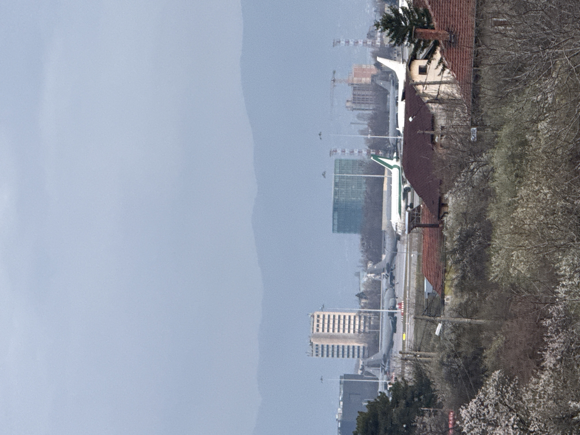 The image presents an aerial view of a cityscape, notably featuring several airplanes positioned near buildings. The presence of distinctive red-tiled roofs suggests a location in Sofia, Bulgaria. The weather appears overcast, casting a diffused light across the scene. Buildings of varying heights and architectural styles punctuate the skyline. Bare trees and early spring blossoms are present. There are no people visible in the image.