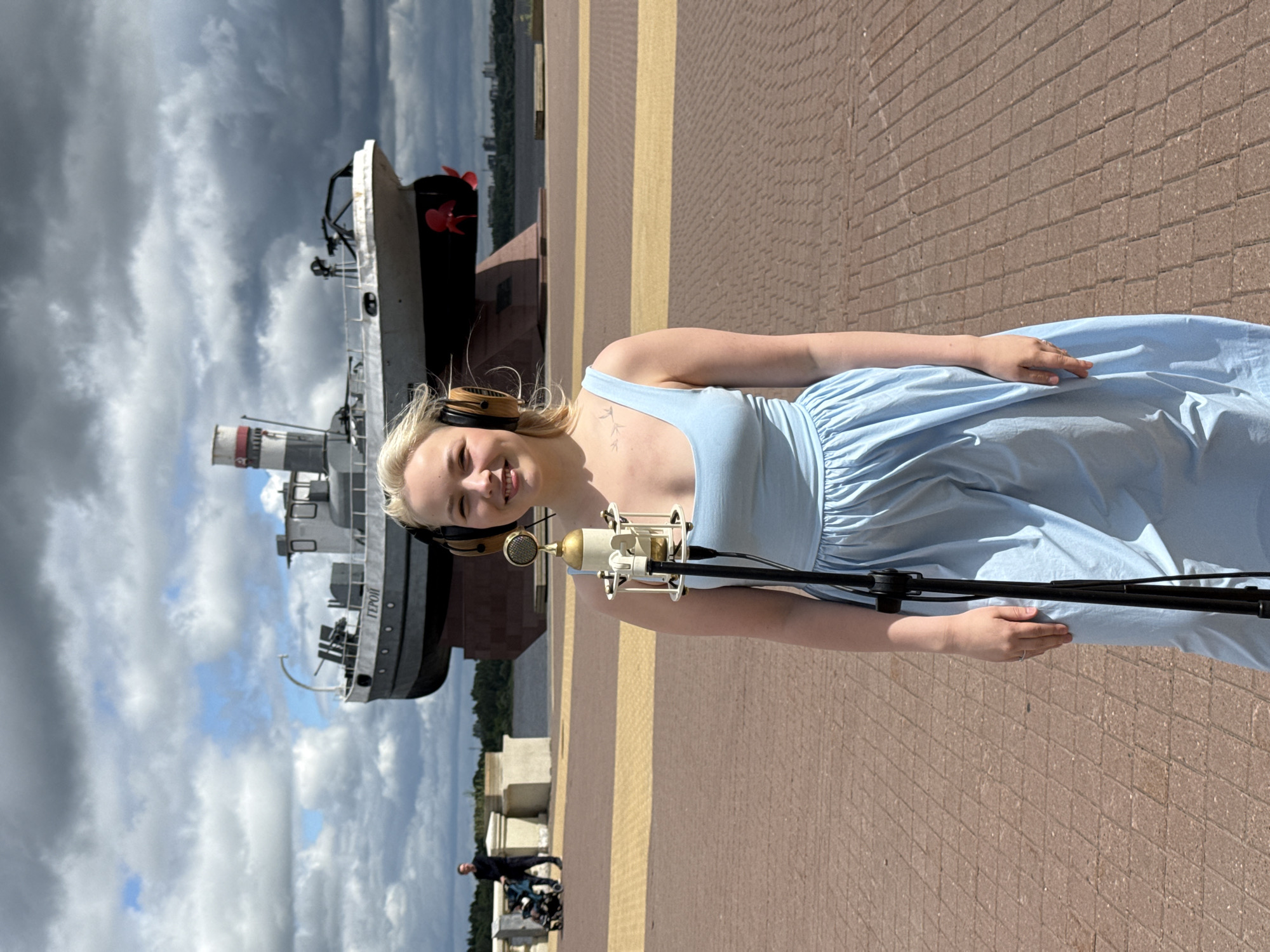 A blonde woman wearing a light blue dress and headphones stands in front of a microphone on a paved promenade. Behind her, a large, dark-colored ship is mounted on a concrete pedestal. The ship has the word "ГЕРОЙ" (Hero) inscribed on its side. In the background, a man pushes a stroller, and further back, a cityscape is visible across a body of water. The sky is cloudy. This scene is located on the Nizhny Novgorod Kremlin embankment, which offers a view of the confluence of the Oka and Volga rivers.