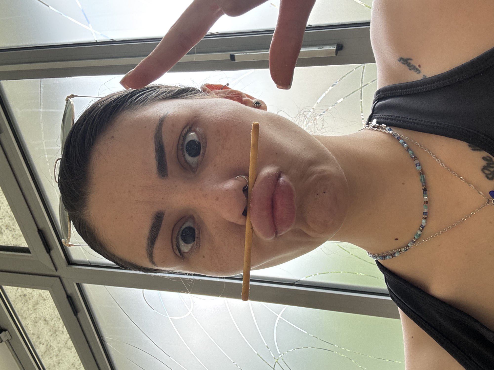 An extreme close-up, inverted portrait captures a person from the shoulders up, posing playfully indoors during the daytime. The subject, appearing to be female, is seen from a low angle, looking directly into the camera with wide, expressive eyes. Her head is tilted, and she balances a thin, light brown stick, resembling a pretzel stick, horizontally between her nose and puckered upper lip.
She wears a black tank top or sports bra. Several accessories adorn her: a beaded necklace in shades of blue and green, a delicate silver chain necklace with a small blue pendant, a nose ring (septum piercing), and earrings (a small stud and a more elaborate ear cuff/multiple piercing visible on her right ear). Sunglasses are pushed up onto her dark hair, which is pulled back from her face.
In the upper part of the frame, two fingers from her right hand are visible, forming a gesture reminiscent of a peace sign. Her skin displays freckles, and her eyebrows are dark and well-defined. Visible tattoos include a small text tattoo on her right shoulder/collarbone area, which appears to be in Thai script, possibly "สิริมงคล" (Sirimongkol - meaning "auspicious" or "blessed"). Another partial text tattoo, with the word "one" visible, is on her side/rib cage area.
The background consists of large, multi-paneled windows or a glass roof structure with grey metal frames, indicating an indoor setting with ample natural light. The glass shows reflections and subtle patterns, suggesting a bright day outside in Sofia, Bulgaria. The overall mood of the image is lighthearted and informal.