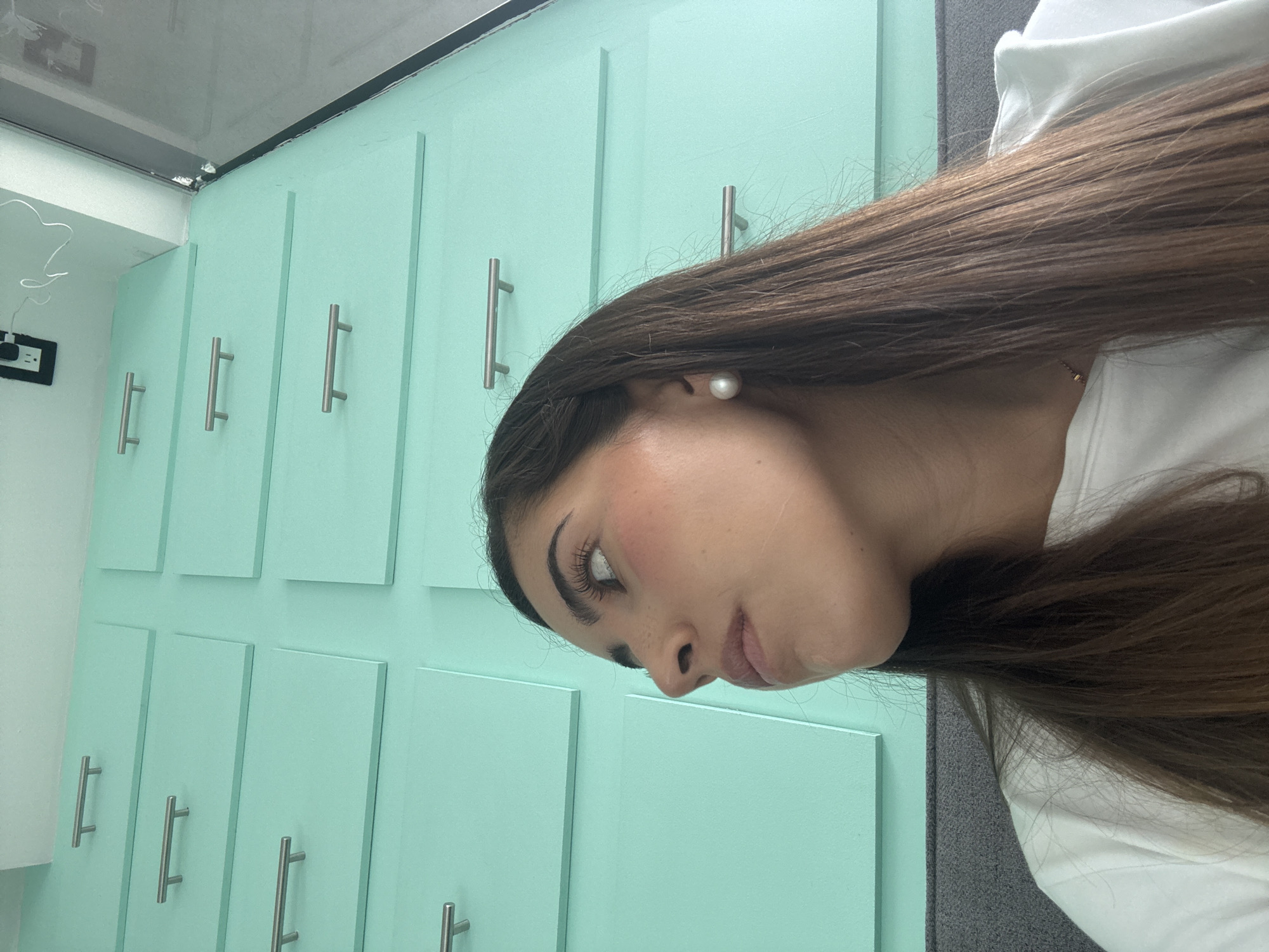 A close-up, rotated view depicts a woman against a backdrop of light teal cabinets with silver hardware. She wears a white top and a pearl earring. Her long brown hair is partially obscuring her face.
There is no specific information to determine if the image was taken in San Bernardino Tlaxcalancingo, Mexico.