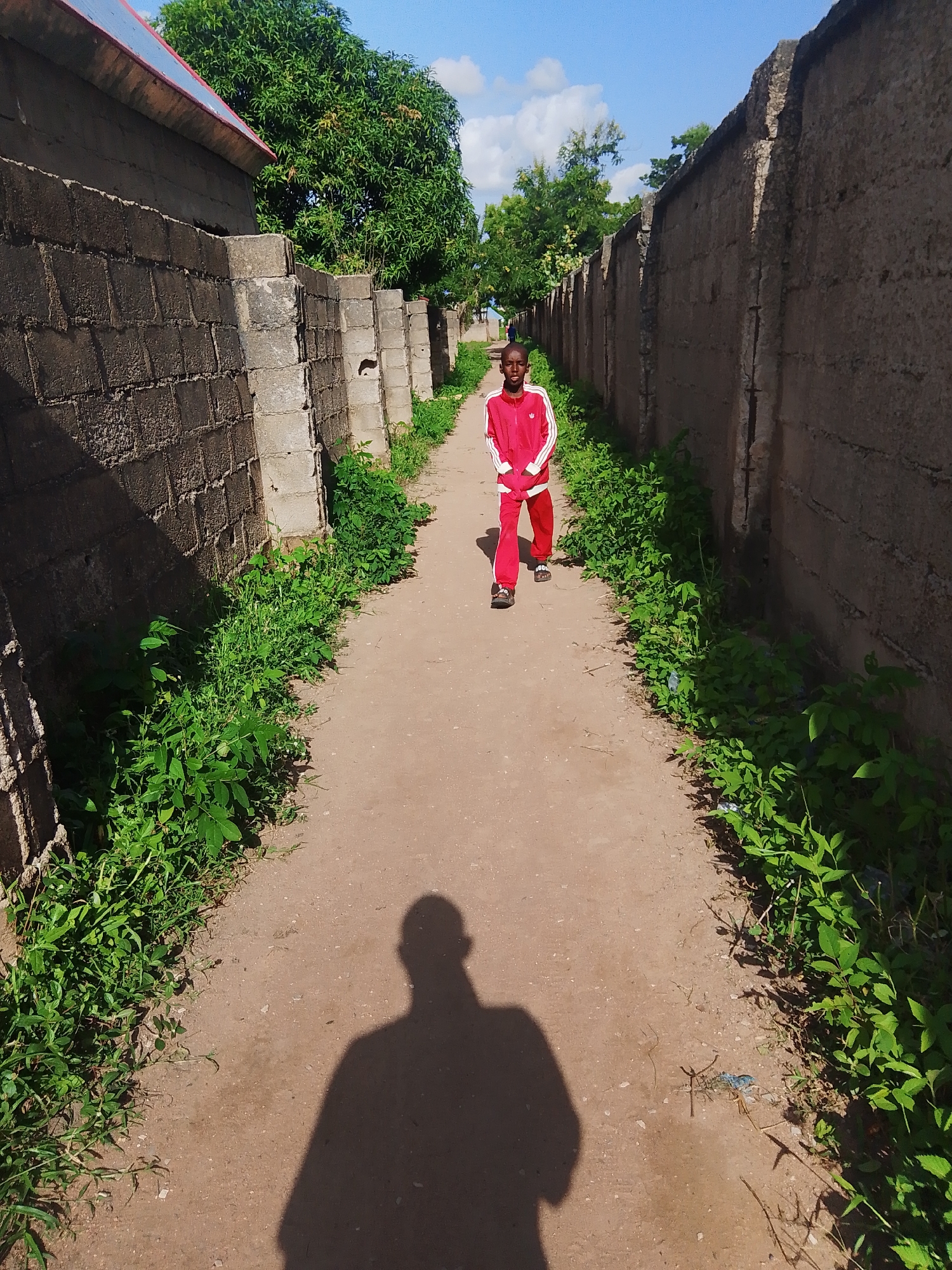 A young boy, likely of African descent, walks down a dusty dirt path in Jalingo, Nigeria. He is wearing a bright red tracksuit with white stripes on the sleeves and down the sides of the pants, and dark sandals. He is the sole person in the foreground of the image, walking towards the viewer.
The path is flanked by tall, rough concrete walls on either side. Lush green vegetation grows abundantly along the base of these walls, some of it reaching onto the path. In the background, trees are visible, and a building with a red roof can be seen to the left. The sky above is a clear blue with scattered white clouds, suggesting fair weather. The sun is high, casting a long shadow of the photographer, and the boy, onto the path. The overall atmosphere is one of a bright, sunny day in a semi-urban or rural African setting. The boy's expression is neutral as he walks. There is no visible text in the image.