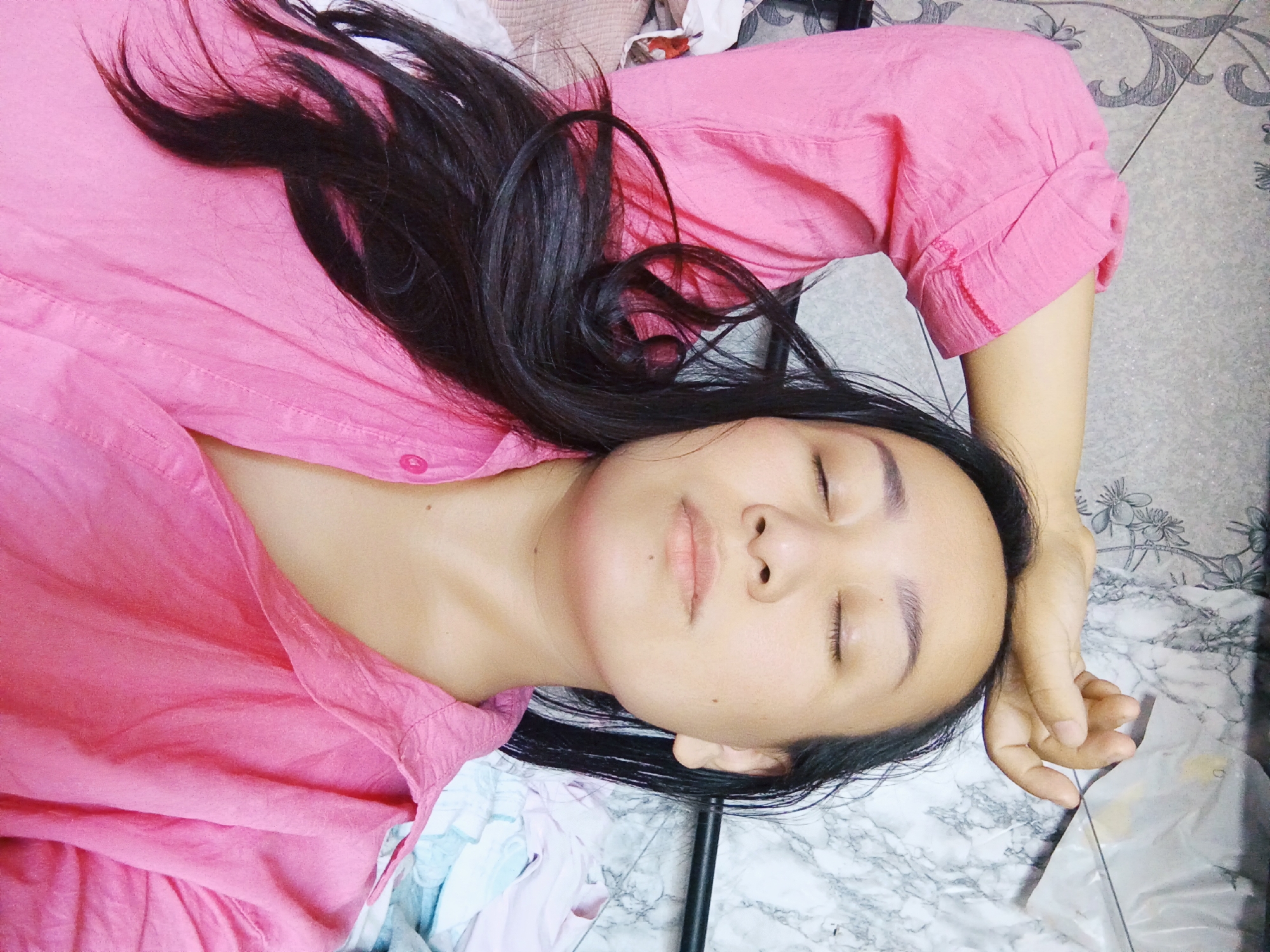An overhead shot shows a woman lying on her back, eyes closed, with her right arm raised and hand resting on her forehead. She is wearing a pink button-down shirt. Her dark hair is spread out around her head. The background appears to be a bed with a patterned headboard and some folded clothes. There is no information about the location in Bishkek City, Kyrgyzstan.