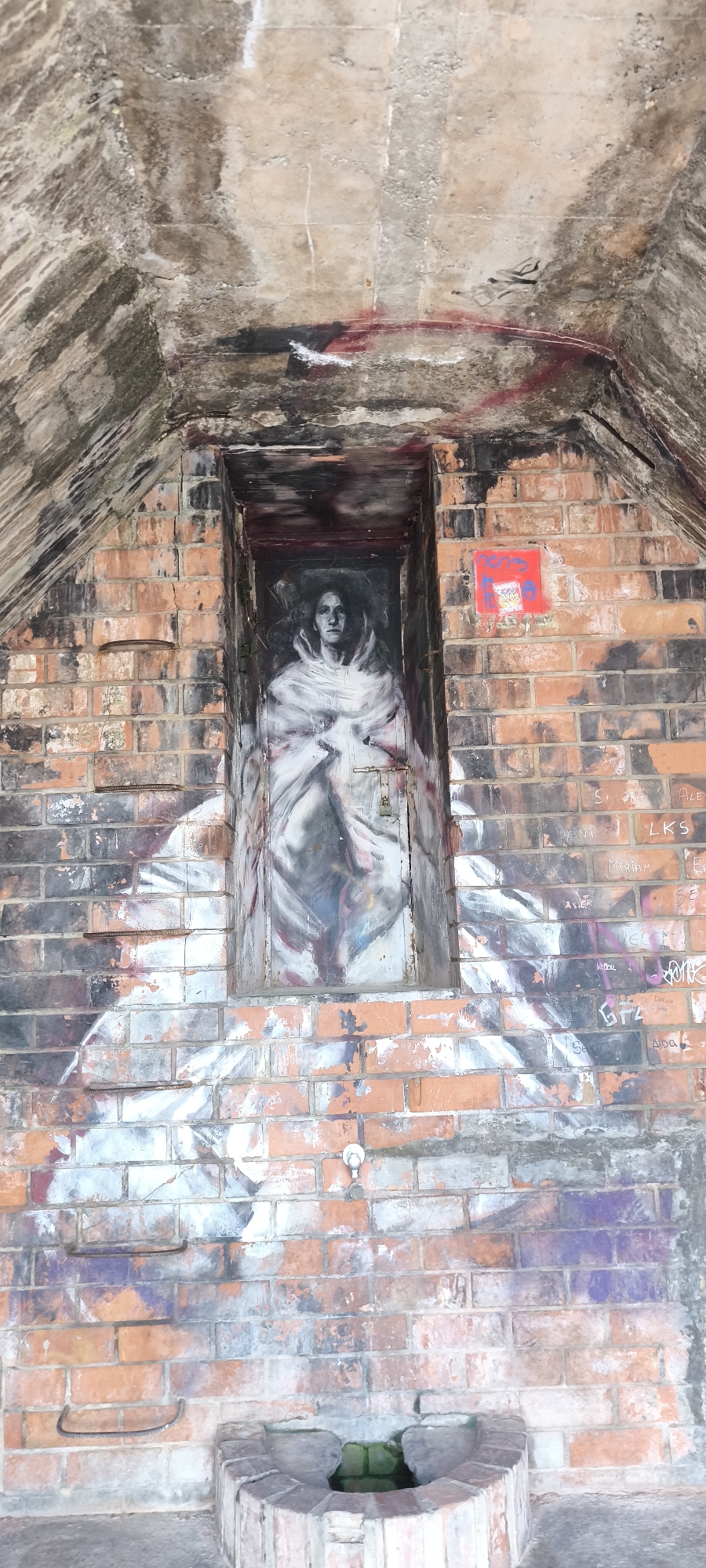 The image shows an outdoor scene in Fanzara, Spain, likely an urban art installation or a derelict structure. Dominating the frame is a weathered brick wall featuring a large, striking mural. The mural depicts a portrait of a woman with a somber expression, rendered in grayscale with subtle hints of color. She is wearing a voluminous white garment that drapes around her shoulders and chest, creating a dramatic effect. The mural is painted within a recessed rectangular opening in the brickwork, resembling a doorway or niche, and is secured by two metal latches and a padlock.
The brick wall itself is aged and shows signs of wear, with some bricks chipped or discolored. Above the mural, the concrete ceiling of an arched structure is visible, stained and marked with graffiti, including red and black spray paint. To the right of the mural, a small red square tag with some writing is affixed to the bricks, and further down, various names and tags are scrawled in graffiti on the wall.
Below the mural, more abstract graffiti in shades of white, gray, and black covers the brickwork, suggesting a continuous artistic theme across the wall. At the very bottom of the image, a circular brick structure is visible, appearing to be a wellhead or a planter, with some green material or water inside. The overall impression is one of an urban environment repurposed for artistic expression, with elements of decay and renewal.