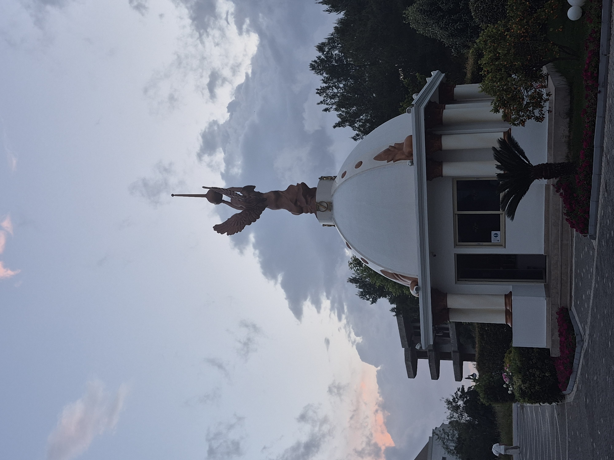 The image shows a statue of a winged figure holding a sphere and a staff, mounted on a building in Alanya, Turkey. The building has a white dome and a facade with columns. A palm tree is visible next to the building, and a garden with flowers lines the pavement. The sky is cloudy.